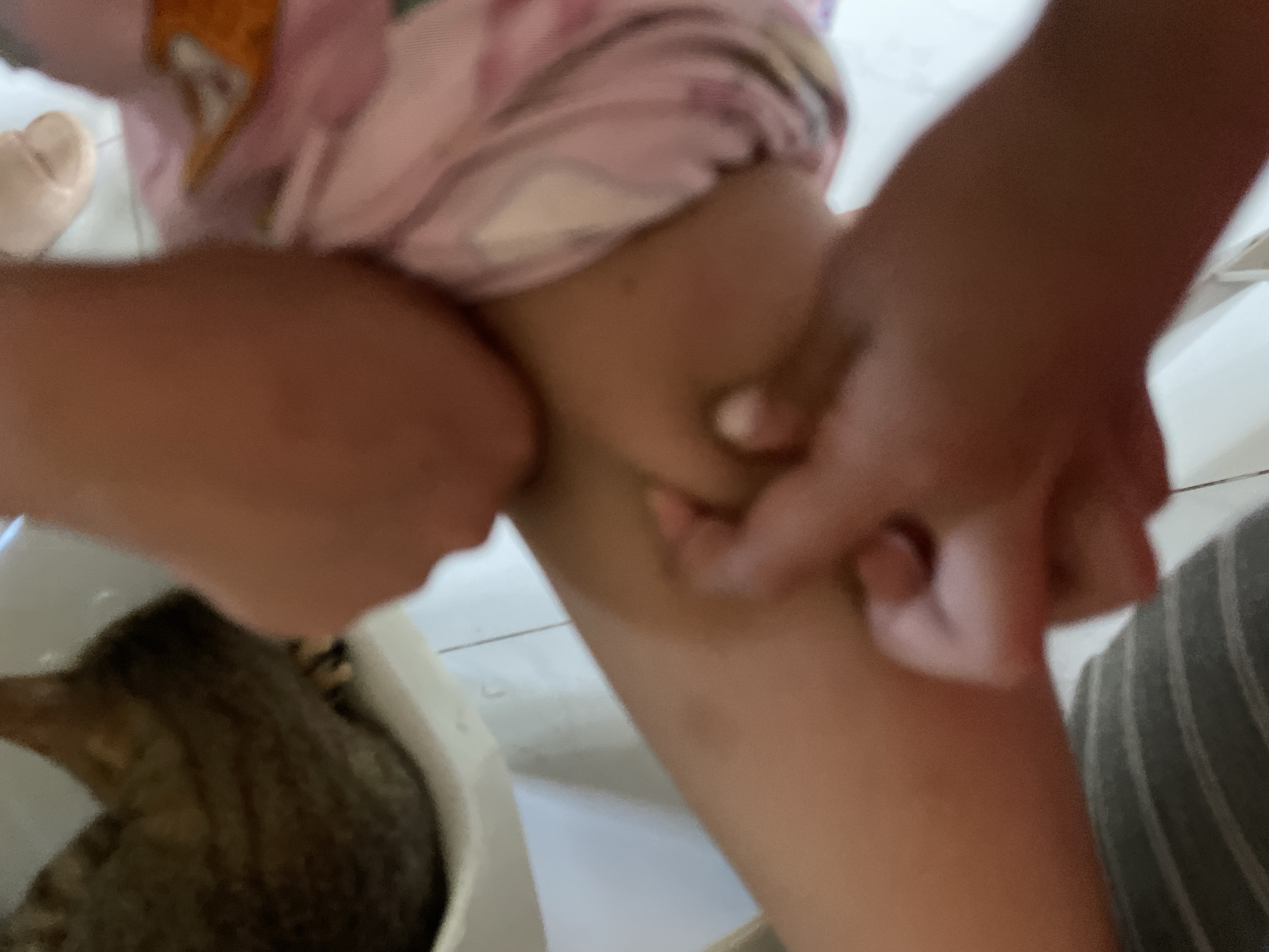 An indoor, blurry close-up photograph captures a person's hands holding or pinching the skin on a leg, while a tabby cat is partially visible in a white container in the lower left. The hands, belonging to a person, are light-skinned and appear to be pressing or gathering the skin on a leg, which is also light brown in tone. The person's upper body is out of focus, but hints of light-colored, possibly pink and patterned clothing are visible. The leg itself shows no distinct features or marks due to the blur. To the left, a cat with brown and black tabby markings is curled up or sitting inside a white bucket or container, its head and back visible. The background consists of a white tiled floor and possibly other blurry indoor elements. The lighting suggests an indoor setting, likely during the day, but no direct light sources are visible. There is no discernible text or clear emotional expression due to the close crop and blurriness. The scene appears to be a casual moment within a domestic environment.