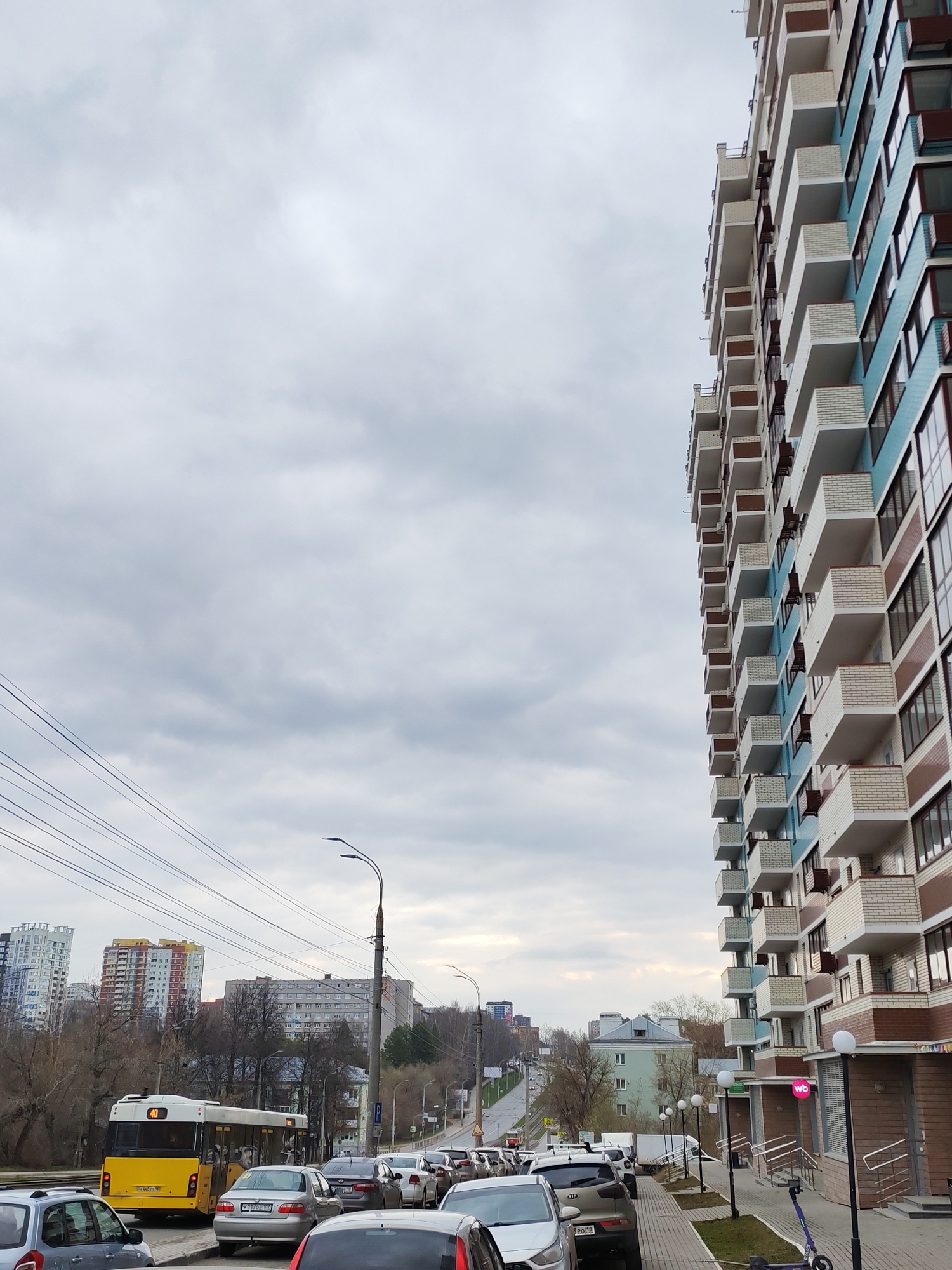 The image depicts a street scene in Izhevsk, Russia, under an overcast sky. A multi-story residential building with protruding balconies is on the right side. The building has a color scheme of white, blue, and brown. The street is lined with parked cars, leading up a slight incline. A yellow bus is on the left side of the street. In the background, other buildings are visible, including a few high-rise structures with colorful facades. Overhead power lines run across the sky. Sidewalks line the street. Streetlights are interspersed along the road.