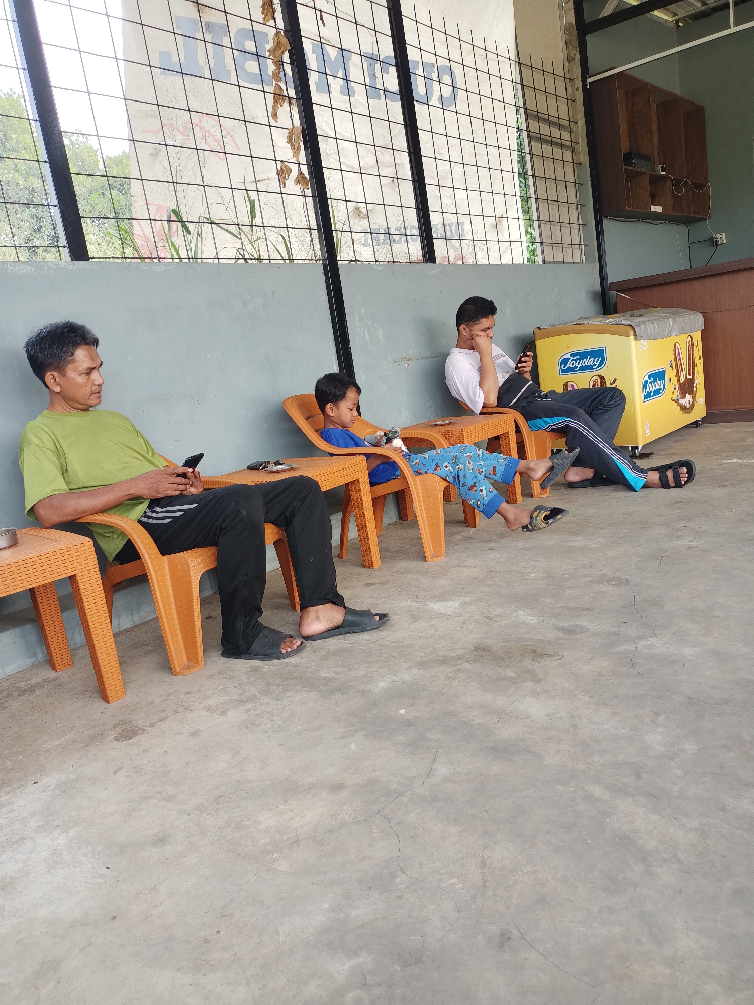In Tajurhalang, Indonesia, three individuals are seated on orange plastic chairs outdoors, in front of a gray wall and behind a metal grate fence. The man on the left, wearing a green shirt and black pants, is holding a smartphone. A child in a blue shirt and patterned pants sits in the middle chair, also looking at a device. To the right, a young man in a white shirt and black pants with blue stripes holds a smartphone and rests his chin on his hand. Adjacent to the chairs is a yellow ice cream freezer, labeled "Joyday." The ground is a concrete surface.