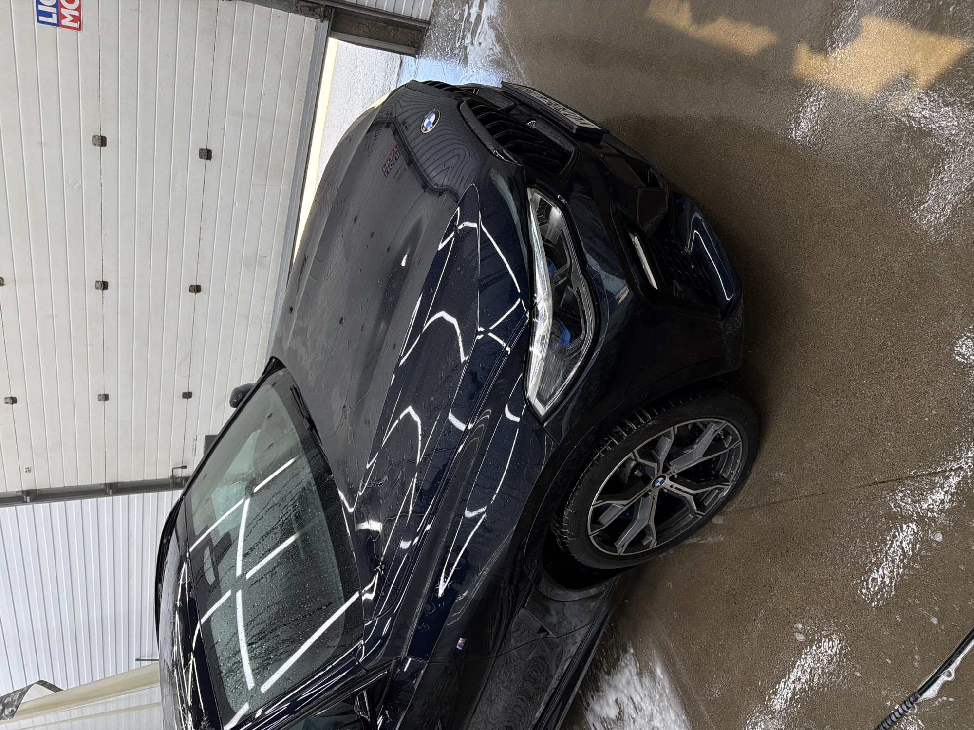 A dark blue BMW SUV is parked on a wet, reflective surface, possibly a car wash bay. The car's hood and roof are wet, with water droplets visible. The car has a large grille and sleek headlights. In the background, a white sectional garage door is visible, with a sign that appears to say "LIQUI MOLY". The ground is covered in water and some foam, indicating recent cleaning. The lighting suggests an indoor or covered environment.
