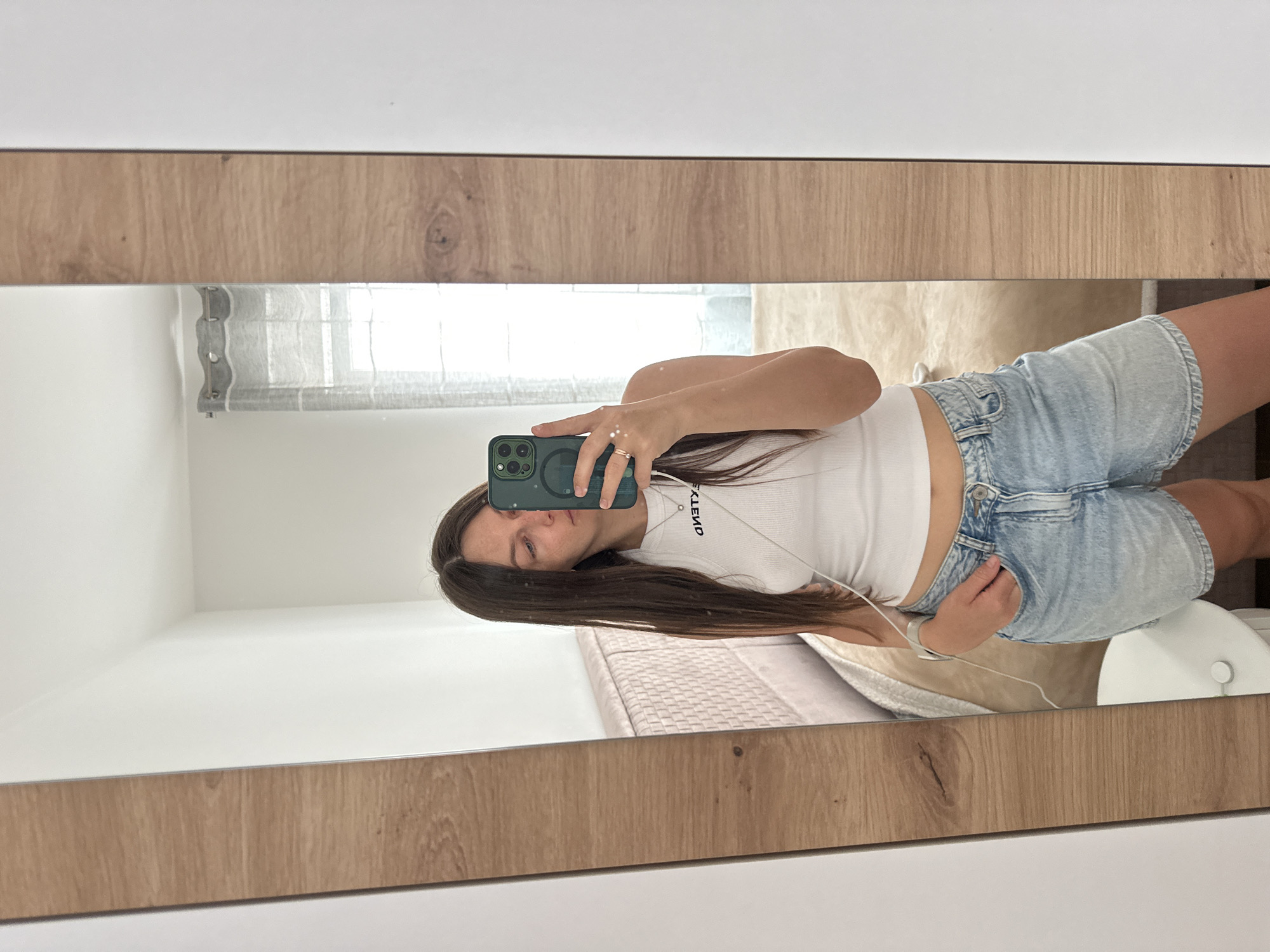A woman is taking a mirror selfie in an indoor setting, presumably a bedroom. The room appears to be located in a residential building in Budva, Montenegro, as the mirror reflects a window with a view typical of the urban landscape found in that area. The woman is wearing a white tank top and denim shorts. She is holding a smartphone with a green case, using it to capture the reflection of herself in the mirror. Her hair is long and brown. She is wearing a ring on her left hand. The mirror has a light wood-tone frame, and it is attached to what appears to be white cabinets or a wall. A bed with a light-colored quilt can be seen in the reflection.