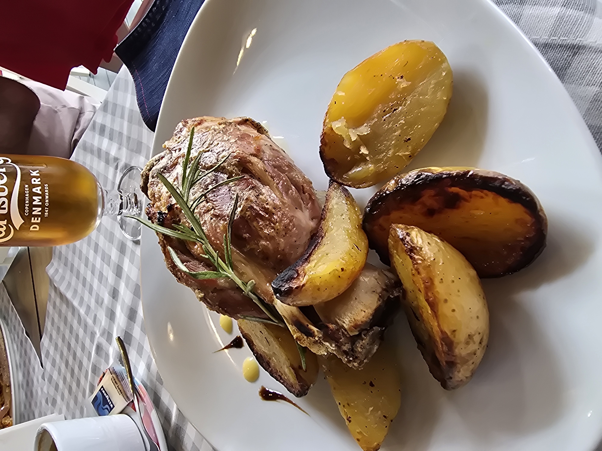 A close-up, eye-level shot captures a white plate with a serving of roasted meat and potatoes. The meat, a substantial piece with visible bone structure and a rosemary sprig on top, is golden brown with hints of charring. Surrounding the meat are several wedges of roasted potatoes, some with crisp, browned edges and others with a softer, glistening interior. A few small dollops of yellow sauce and a drizzle of dark sauce are artfully placed on the plate. The plate rests on a grey and white checkered tablecloth. In the background, a glass of amber liquid, likely beer, with a Carlsberg logo is visible, along with a white cup and a small packet of Lavazza coffee. The scene appears to be set outdoors, possibly at a restaurant or cafe.