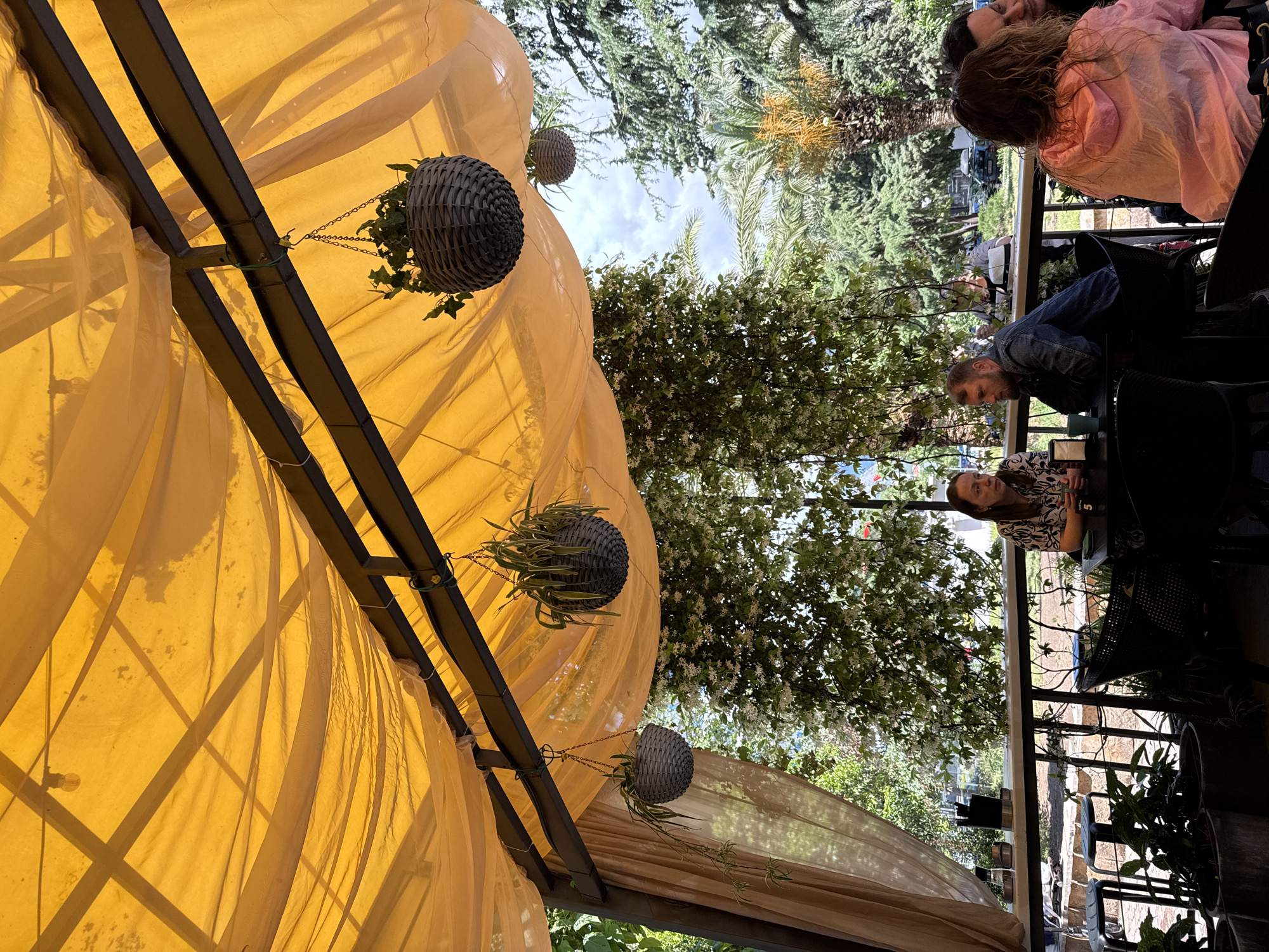 The image captures a scene at a cafe in Bar, Montenegro. The foreground features a yellow fabric canopy, partially obscuring the view. Three hanging planters with trailing greenery are suspended from the canopy's metal frame.
Beyond the canopy, a group of people are seated at tables. Two women are visible: one wearing a black and white patterned shirt and another in a pink hooded jacket. A man in a denim jacket is also present. They are seated around tables with drinks, suggesting a casual social interaction.
The background displays a mix of urban elements and natural scenery. Trees and other foliage are visible, as well as parts of buildings or structures in the distance. The sky is partly visible through the trees.