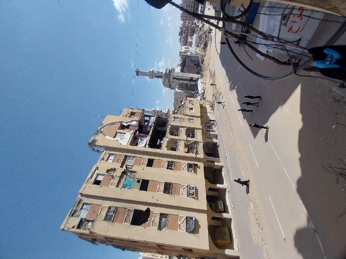 An aerial-perspective image depicts a scene of extensive urban destruction in Khan Yunis, Palestinian Territory, under a clear blue sky with scattered clouds. In the foreground and midground, a paved road with white lane markings spans horizontally.
To the left of the road stands a multi-story residential building, approximately seven to eight stories tall, constructed with a beige facade. This building exhibits severe damage, including large holes penetrating its structure, missing sections of walls and balconies, exposed interior rooms, and visible impact marks consistent with projectile damage across its exterior. A light-colored van is parked beneath the arched ground-floor section of this damaged building.
In the background, beyond the road and to the right of the damaged apartment building, the partially collapsed structure of a mosque is visible. Its minaret remains standing, although the main prayer hall and dome appear to be heavily damaged or destroyed. Surrounding the mosque and extending further into the background is a large expanse of rubble and debris from numerous other collapsed buildings, indicating widespread destruction.
On the paved road, a group of five individuals, dressed in dark clothing (possibly uniforms), stand together on the right side. Another individual, clad in a long dark garment, walks across the road from left to right in the midground. Two additional small figures are discernible amidst the distant rubble. Shadows from people and unseen structures are cast across the road, suggesting bright daylight. The lower right foreground is partially obscured by dark cables and a blurry structure, behind which a white surface with partial Arabic text "جراح" is visible.