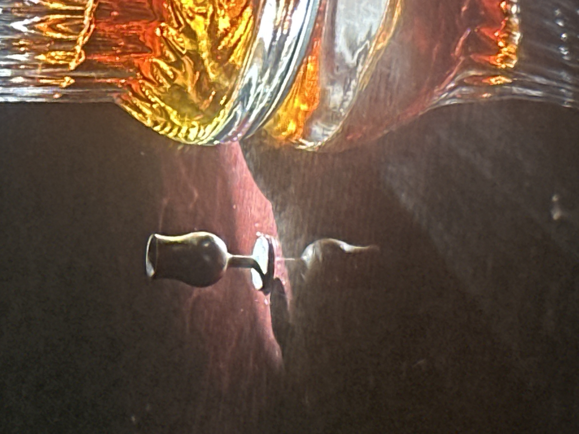 The image shows a close-up of a table with a small, dark glass on the left, casting a shadow. To the right, a large, ornate glass decanter filled with amber liquid is partially visible, its surface reflecting warm light. The decanter has vertical, ribbed detailing and a textured pattern on its lower half. The background is dark and indistinct, suggesting a dimly lit environment. The scene is a still life composition. There is no information within the image to indicate a specific location in the city of Lobacheve, Ukraine.