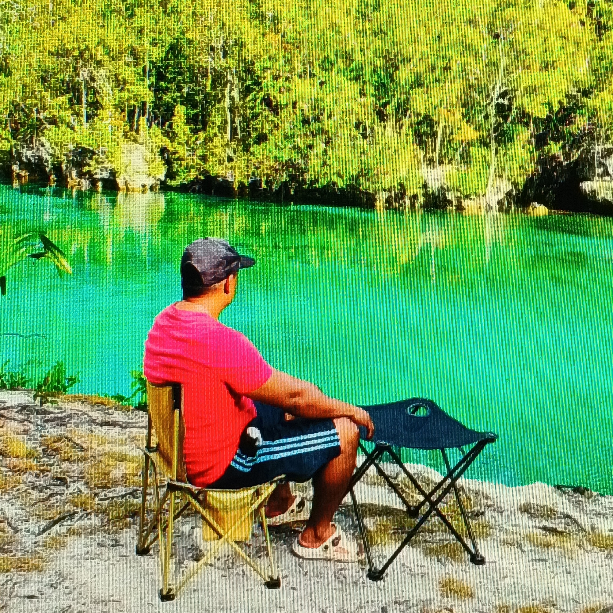 A person is sitting on a folding chair facing away from the camera, looking out at a vibrant turquoise body of water bordered by lush green foliage. The person is wearing a red t-shirt, dark shorts with white stripes, a grey baseball cap, and light-colored sandals. Next to the person, to their right, is a black folding table. The ground appears to be sandy with some dry vegetation. The water reflects the green trees and sky. The scene is tranquil and appears to be a natural setting. Without further information or visual cues within the image, it is impossible to definitively place this scene in Ende, Indonesia.