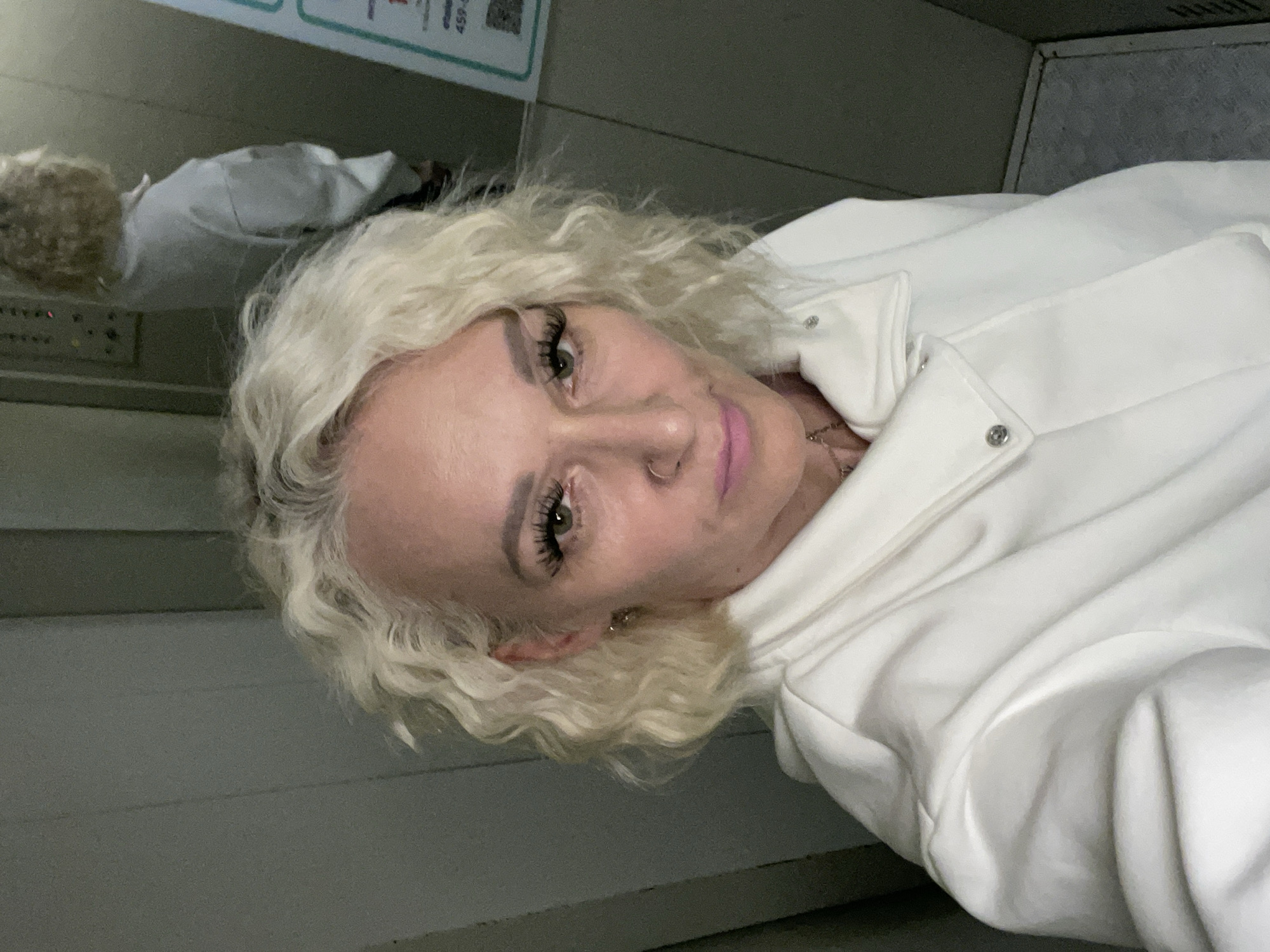 A woman with blonde curly hair and noticeable makeup takes a selfie in an elevator. She is wearing a white jacket with silver snaps. In the background, another person with blonde curly hair is visible, facing away from the camera, also wearing a white shirt. The elevator walls appear to be grey. A sign with text and a QR code is partially visible to the right of the frame. The floor of the elevator is visible at the bottom, made of grey metal grating. The overall lighting is dim.