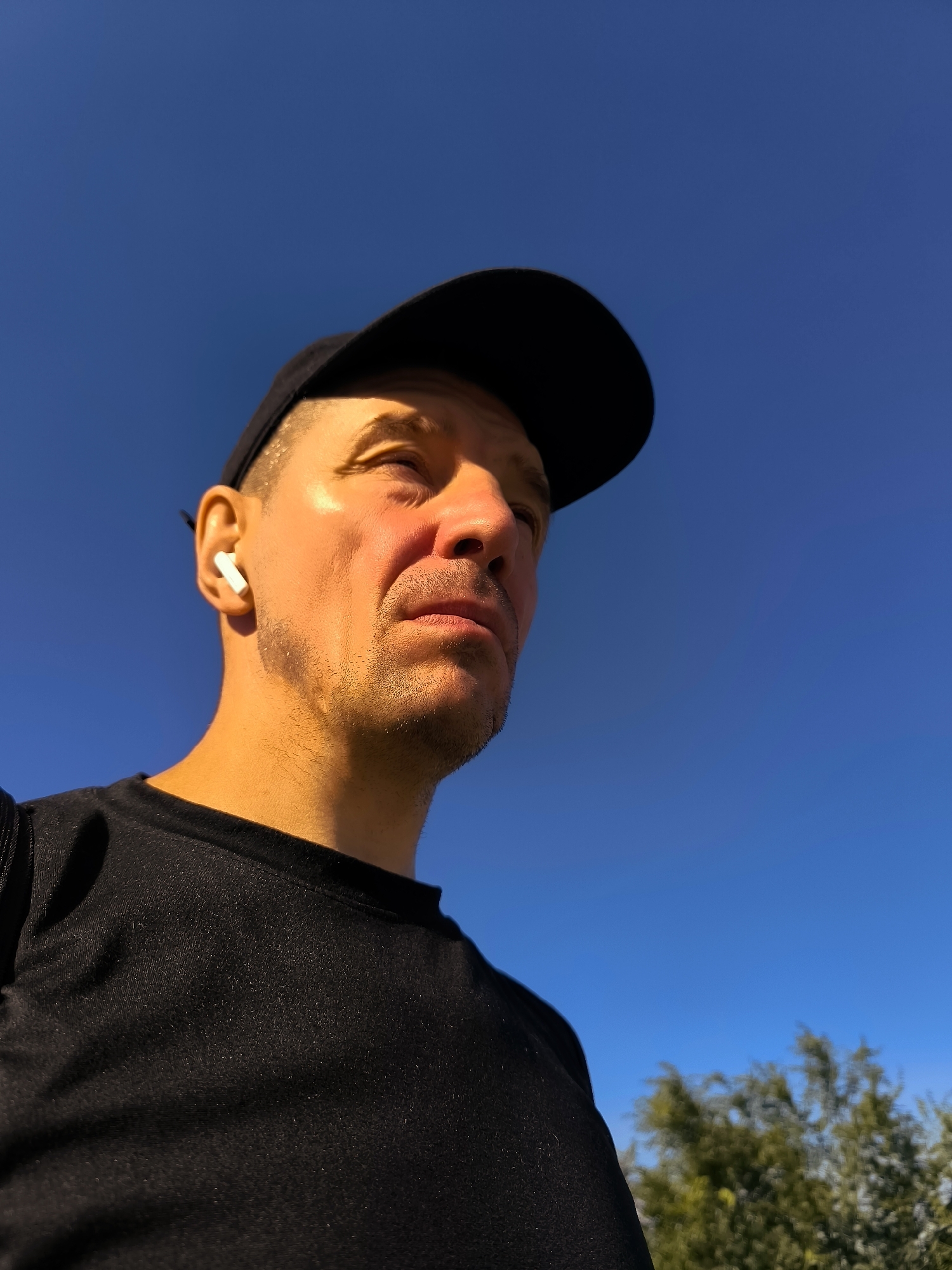 A low-angle medium shot shows a man wearing a black baseball cap and a black t-shirt against a clear blue sky. He has short, stubble-like facial hair and is wearing white wireless earbuds in his left ear. Sunlight illuminates his face from the right, casting a shadow from the cap over the top of his head. In the background, the top of a leafy green tree is visible on the right side.