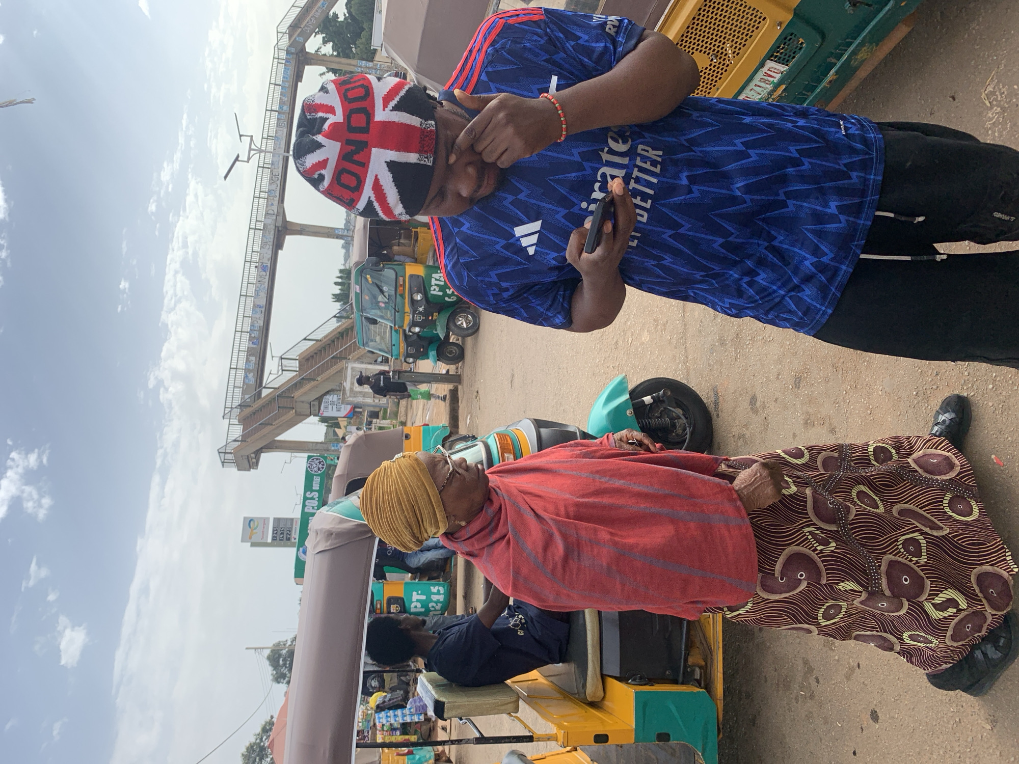 In Gura Top, Nigeria, a man wearing a blue and white Union Jack-patterned beanie and a blue Arsenal football jersey is standing and looking at his phone. To his left, an elderly woman wearing a yellow headscarf and a red shawl over a patterned skirt is standing and looking to her right. They are both standing on a dirt road next to several auto-rickshaws. In the background, there is a pedestrian overpass, a Total gas station, and other vehicles. The sky is cloudy.