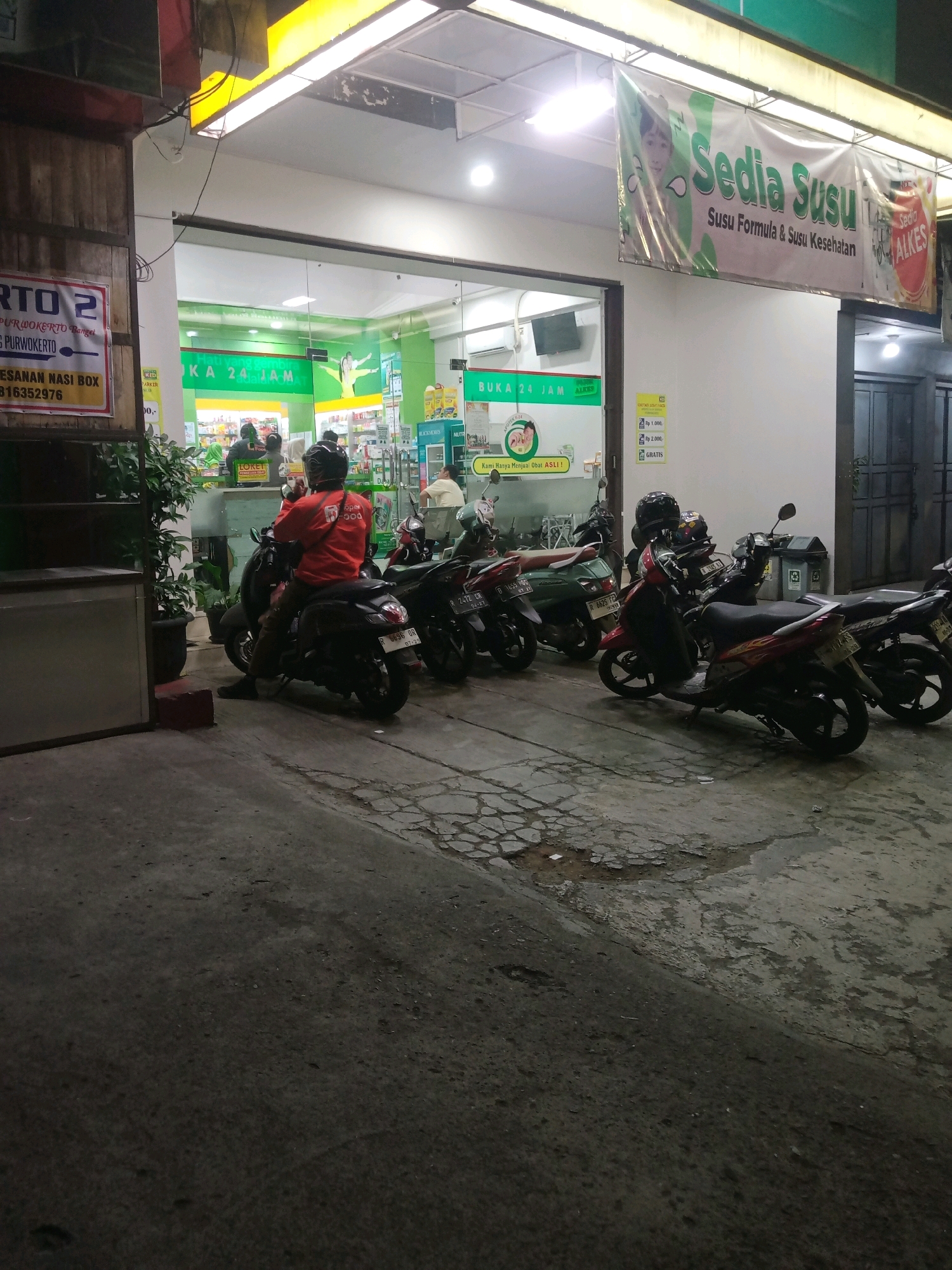 The image depicts the exterior of a brightly lit pharmacy or convenience store in Purwokerto, Indonesia, at night. The store's interior is visible through large glass doors and windows, revealing aisles stocked with products and a counter area where a few people are present. A banner hangs above the entrance, proclaiming "Sedia Susu" (Milk Available) and "Susu Formula & Susu Kesehatan" (Formula Milk & Health Milk), along with images of children and milk products. A sign on the building indicates "BUKA 24 JAM" (Open 24 Hours).
Outside the store, several motorcycles are parked on the pavement. One person, wearing a bright orange jacket with "open FOOD" printed on it, is seated on a dark-colored scooter, appearing to be a delivery rider. Other individuals are also visible near the motorcycles and in the background within the store. The ground in front of the store is paved with concrete and asphalt, showing some signs of wear and tear. A wooden structure with shelves is partially visible on the left side of the image, along with a potted plant. The overall atmosphere suggests a busy commercial area during nighttime operations.