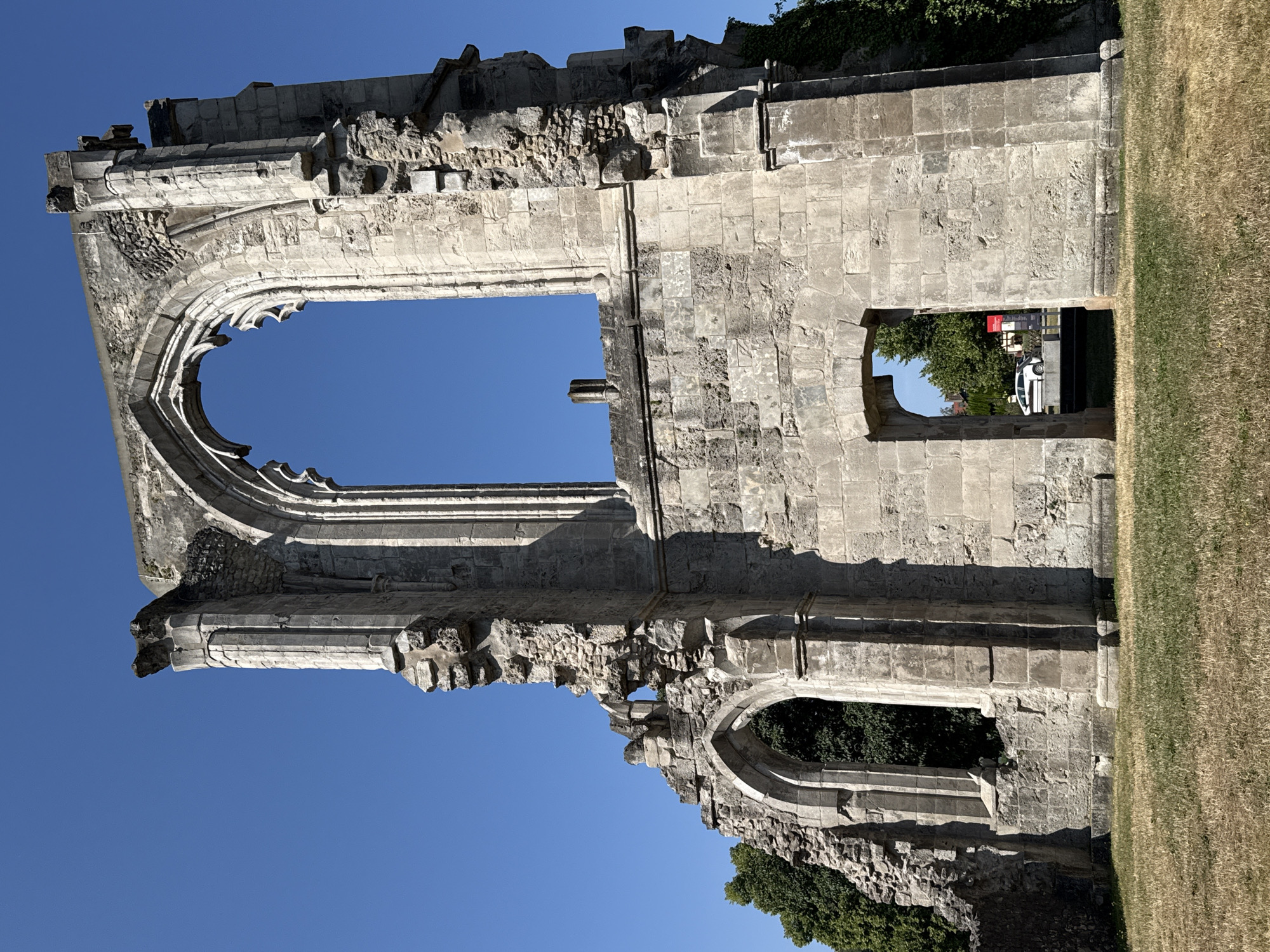 This is a full-shot, eye-level, outdoor photograph of the exterior ruins of a stone building, likely a church or abbey, located in Walkenried, Germany. The ruins are predominantly gray stone, with some sections appearing weathered and incomplete. The architecture features Gothic arches and buttresses. In the background, through the arched openings of the ruins, there is greenery from trees and a clear blue sky. To the right of the ruins, some ivy is growing on the stone. In the far background, a white car is visible parked near a building with a red sign. The ground in front of the ruins is covered with dry, yellow-brown grass. The lighting suggests it is a bright, sunny day.