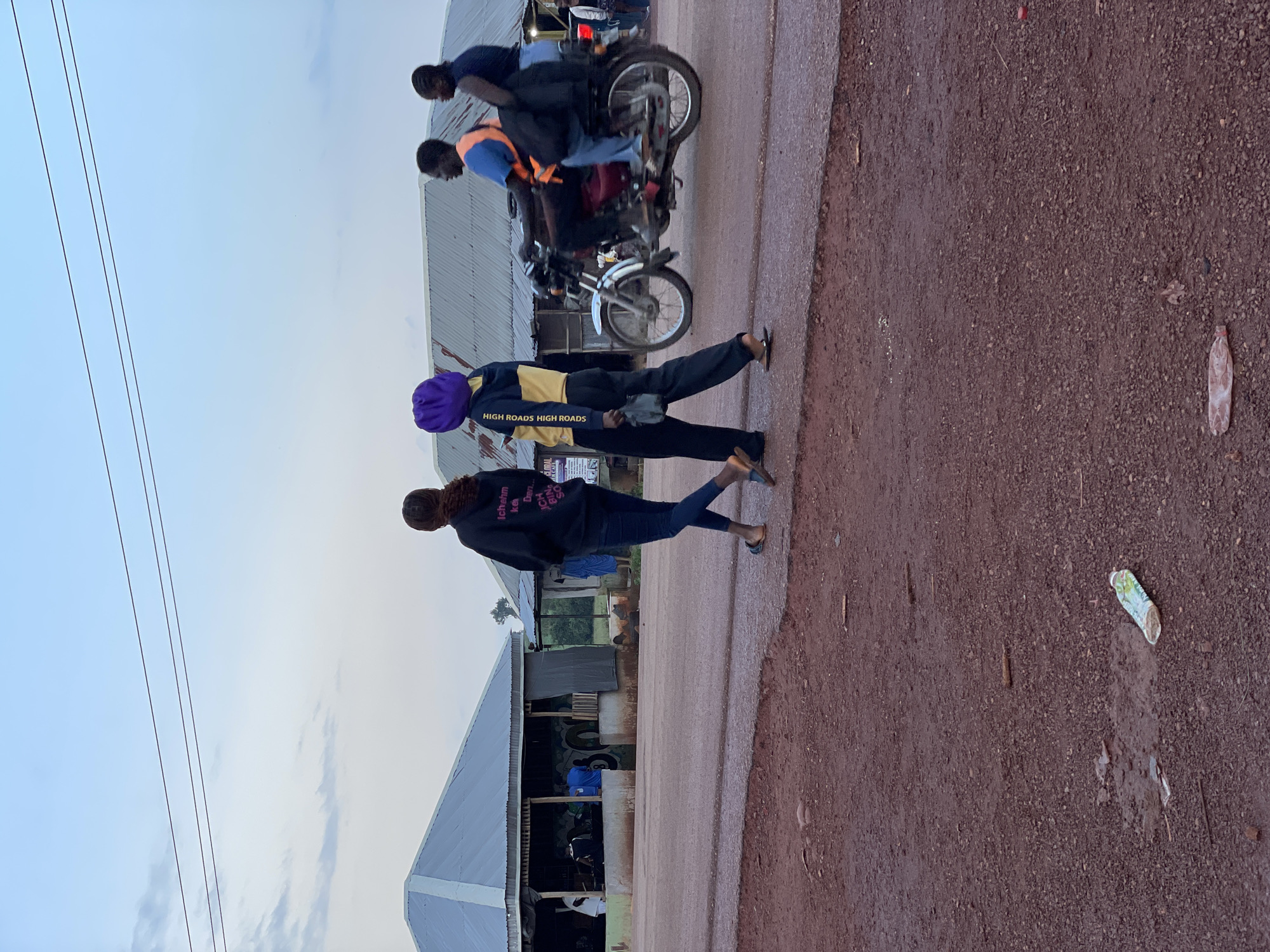 In Durimi, Nigeria, a dirt road stretches across the frame, bordered by buildings with corrugated metal roofs. Two women are seen walking away from the camera on the right side of the road. The woman closer to the foreground wears a dark hoodie and blue jeans, while the woman beside her is dressed in a yellow and black hoodie with a purple headscarf and dark pants. Further down the road, a motorcycle carries two passengers: a man driving and a woman seated behind him. In the background, the buildings appear to be shops or residences. The sky above is overcast with a few wisps of clouds. The foreground of the image shows a patch of red dirt and gravel, with some scattered debris.