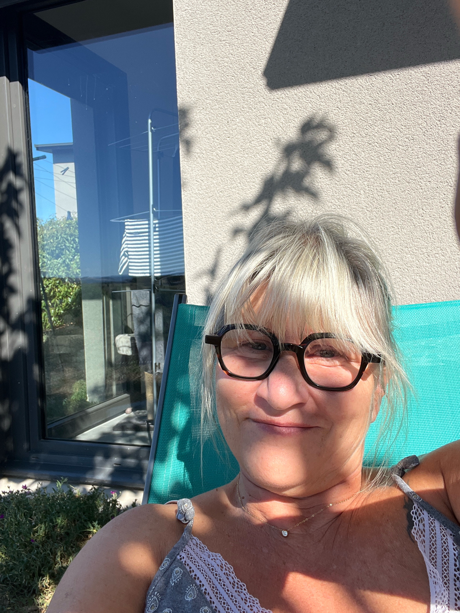 The image shows a medium shot of a middle-aged woman with blonde hair and glasses sitting outdoors on a teal lounge chair. She is wearing a grey patterned top with lace trim and a delicate necklace. The background features a modern building with large windows, a wall with a textured finish, and shadows cast by foliage. The woman is looking at the camera and smiling. The scene appears to be a residential backyard in Chambœuf, France, on a sunny day.