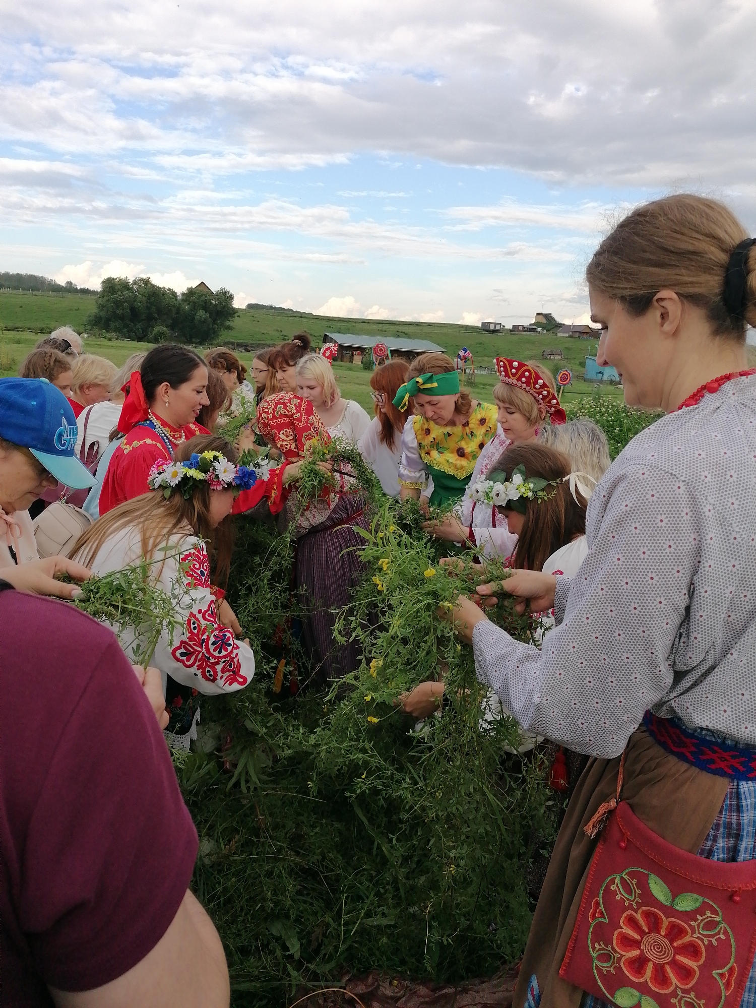A group of people, primarily women and girls, are gathered outdoors in a grassy field under a cloudy sky. They are dressed in traditional clothing, with several wearing embroidered tops and headwear, including flower wreaths. The individuals appear to be engaging in an activity involving green foliage and possibly making wreaths or similar crafts. In the background, there are some scattered buildings and trees. The scene suggests a cultural or celebratory event taking place in a rural setting.