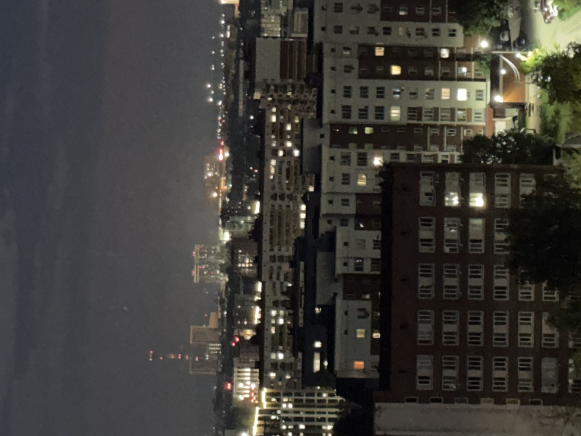 The image shows a nighttime aerial view of a residential area in Izhevsk, Russia. Several multi-story apartment buildings are visible, with many windows illuminated, indicating that the apartments are occupied. In the distance, a tall radio tower with red lights is prominent, along with other buildings and city lights. The sky is dark and overcast. The foreground features the tops of trees and some greenery, suggesting a somewhat built-up urban environment with pockets of nature. The overall scene depicts a typical urban landscape at night.