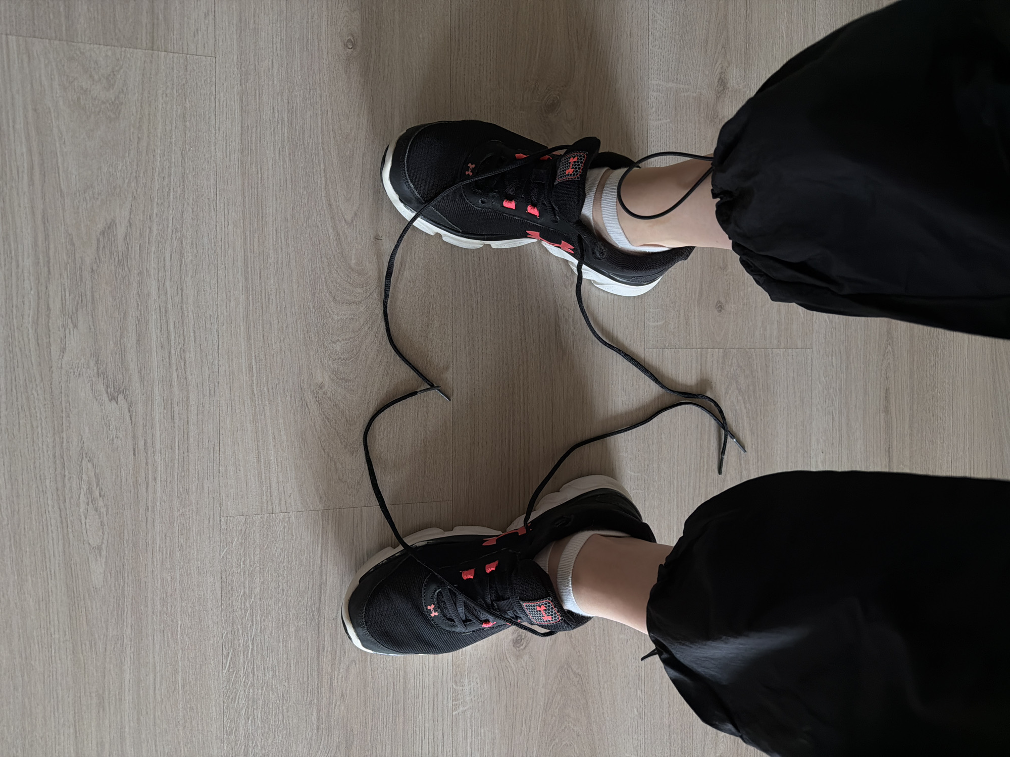 The image shows a top-down view of a person's legs and feet wearing black and coral colored sneakers with untied laces. The person is also wearing white socks and black athletic pants. The floor is light brown wood-patterned laminate. The location is not identifiable as Izhevsk, Russia, based on the provided image.