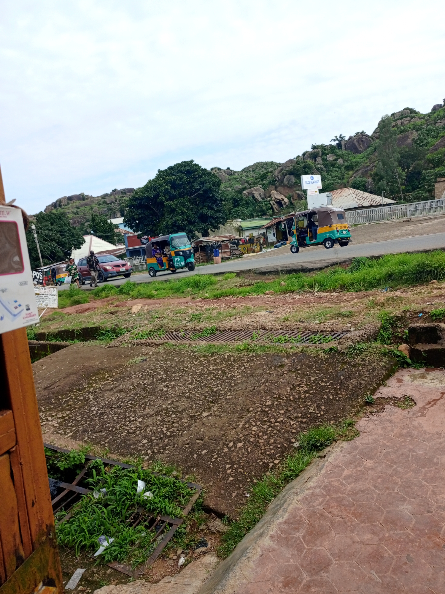 The image depicts a street scene on Rukuba Road, Nigeria. A two-lane asphalt road runs horizontally across the frame, with lush green grass and vegetation bordering it. In the foreground, a concrete drainage grate filled with weeds and some white debris is visible. Beyond the road, there are several buildings and structures, some appearing to be shops or small businesses, with corrugated metal roofs. Rocky hills covered in greenery form the backdrop of the scene.
Several vehicles are present on the road. On the right side, a turquoise and brown auto-rickshaw, commonly known as a "Keke Napep" in Nigeria, is traveling towards the left. A person is visible in the driver's seat. Further down the road, another similar auto-rickshaw, this one primarily yellow, is also in motion. A red car is driving away from the viewer towards the left, and a person in a patterned dress and a man in a dark uniform are walking across the road in front of it. Another auto-rickshaw is parked on the left side of the road. A large white sign with blue lettering is mounted on a pole near the buildings. The sky is overcast with a light gray cloud cover.
