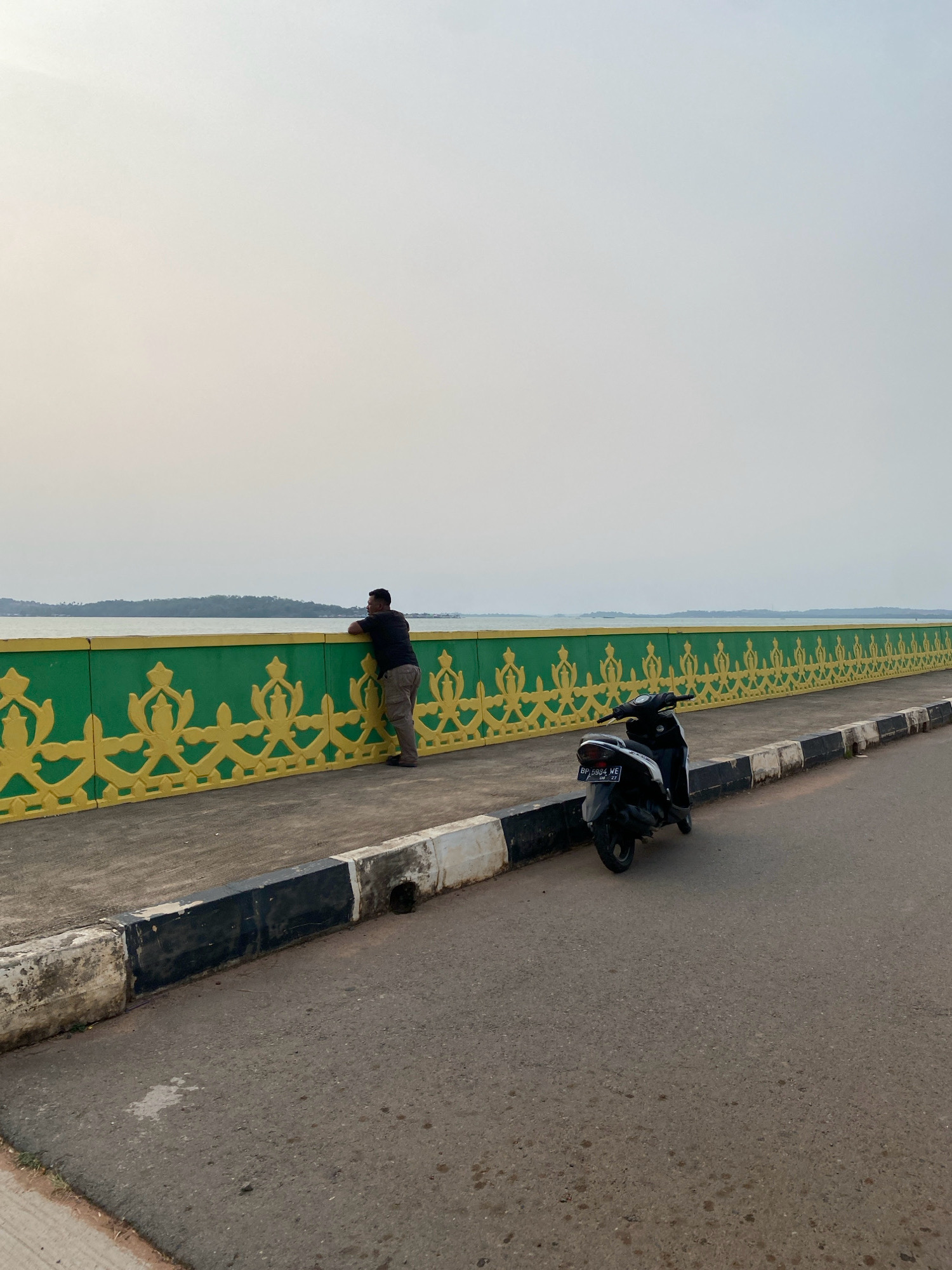 A lone man stands on a pedestrian walkway, leaning over a green and yellow decorative barrier, looking out at a body of water. His posture suggests contemplation or observation. A black scooter is parked on the walkway to his right. The walkway is bordered by a road with a striped curb. The sky is overcast and the light is soft, indicating either early morning or late afternoon. The scene appears to be along the waterfront of Tanjung Pinang, Indonesia, a city known for its coastal scenery and historical sites. The barrier design is typical of local decorative elements found in public spaces, possibly a bridge or promenade.