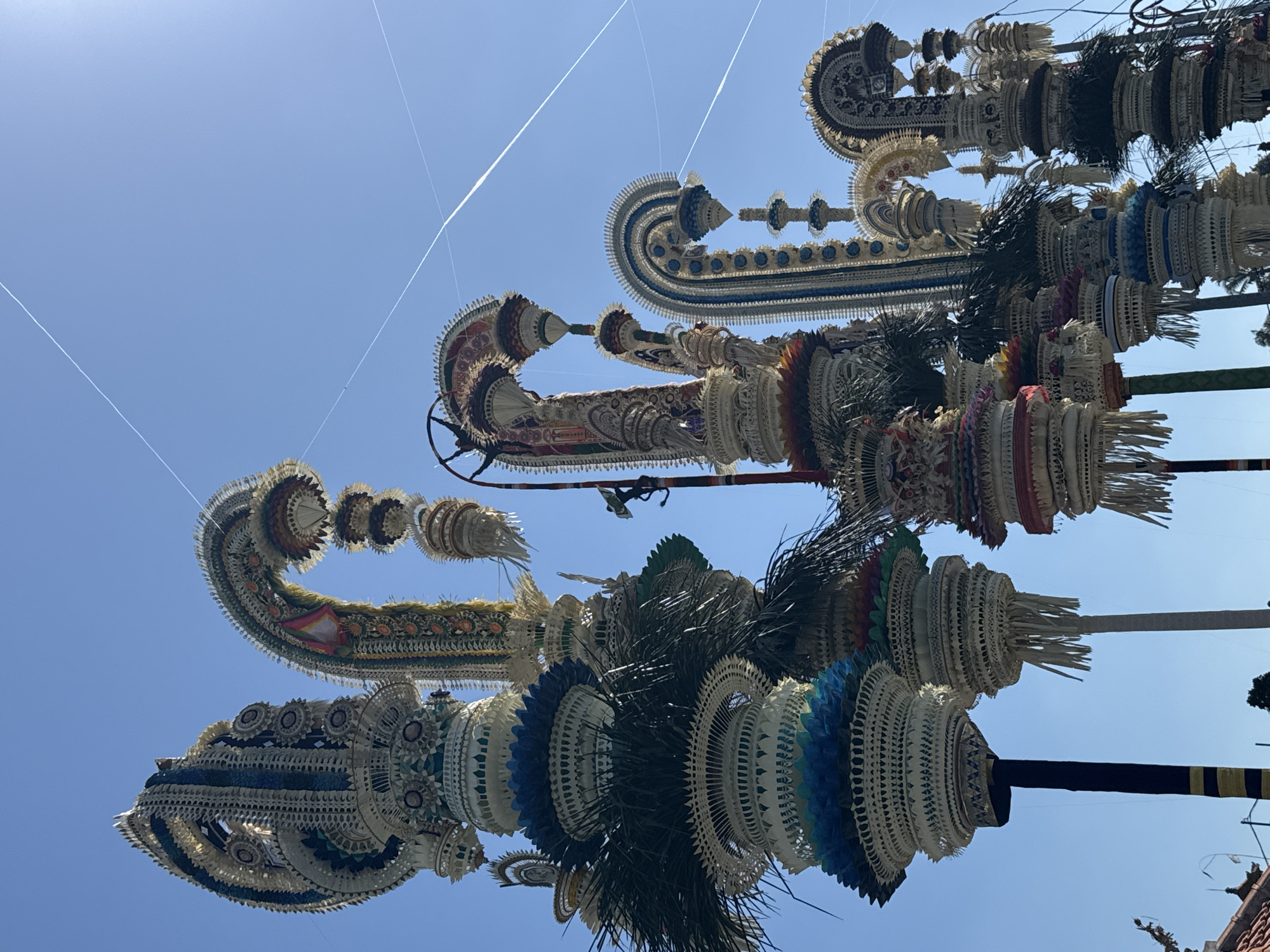 The photograph captures a collection of towering, intricately decorated traditional Balinese structures, known as "penjor," set against a clear blue sky. These elaborate bamboo poles are adorned with woven palm leaves, colorful fabric, and ornate carvings, reaching upwards in curved and tiered formations. The penjor vary in height and design, creating a dynamic visual composition. In the foreground, some of the lower sections of the penjor display vibrant blue and green accents, layered with palm fronds. Towards the background, the structures continue to ascend, their delicate details visible against the bright sky. Faint white contrails from airplanes are visible in the upper portion of the image. The scene appears to be outdoors, possibly during a festival or religious ceremony, given the grandeur and artistry of the penjor. While the specific location is given as the city of Mengwi, Indonesia, no people or explicit actions are captured in the frame, focusing entirely on the impressive architectural and decorative elements of the penjor themselves. The overall impression is one of cultural richness and meticulous craftsmanship.