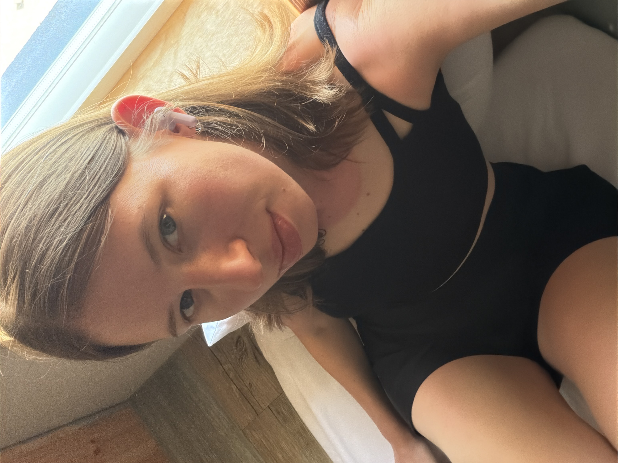A young woman is lying on a bed, taking a selfie. She has light brown hair and blue eyes. She is wearing a black sports bra and black shorts. She has a light pink earbud in her right ear. The room appears to be a hotel room, with wooden walls and a large window. The lighting is natural, coming from the window. There is a pillow behind her head, and a white sheet on the bed. The overall impression is one of relaxation and comfort. The image was taken at a high angle, looking down at the woman.