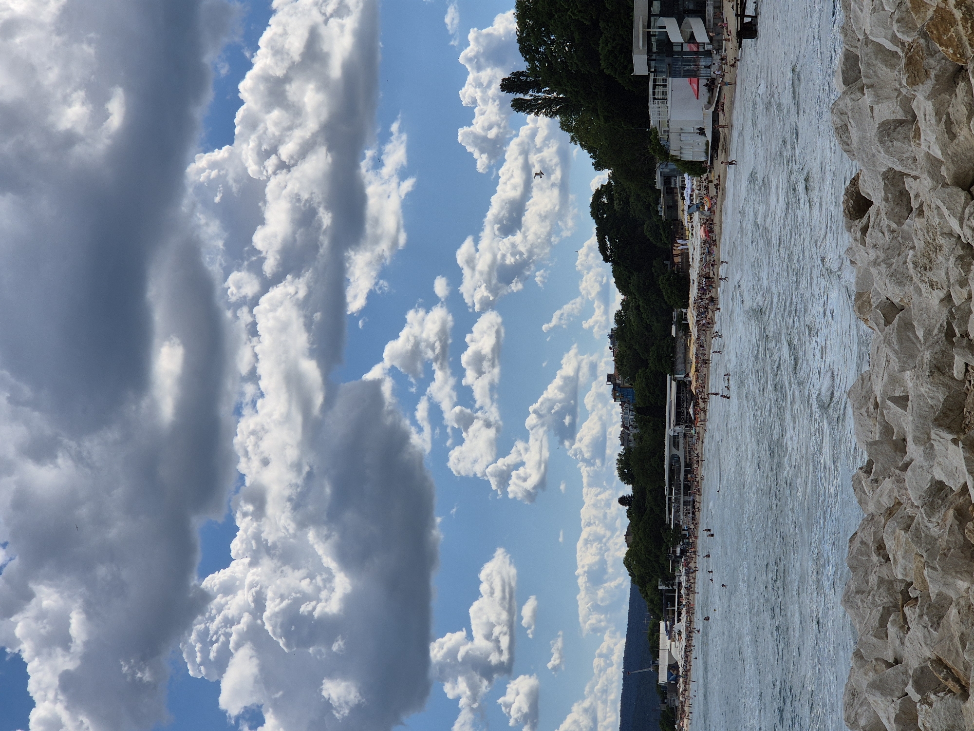 The image shows a crowded beach with many people sunbathing and swimming in the ocean. The water is choppy with small waves. In the background, there are trees, buildings, and a cloudy sky with some birds flying. In the foreground, there are large rocks.