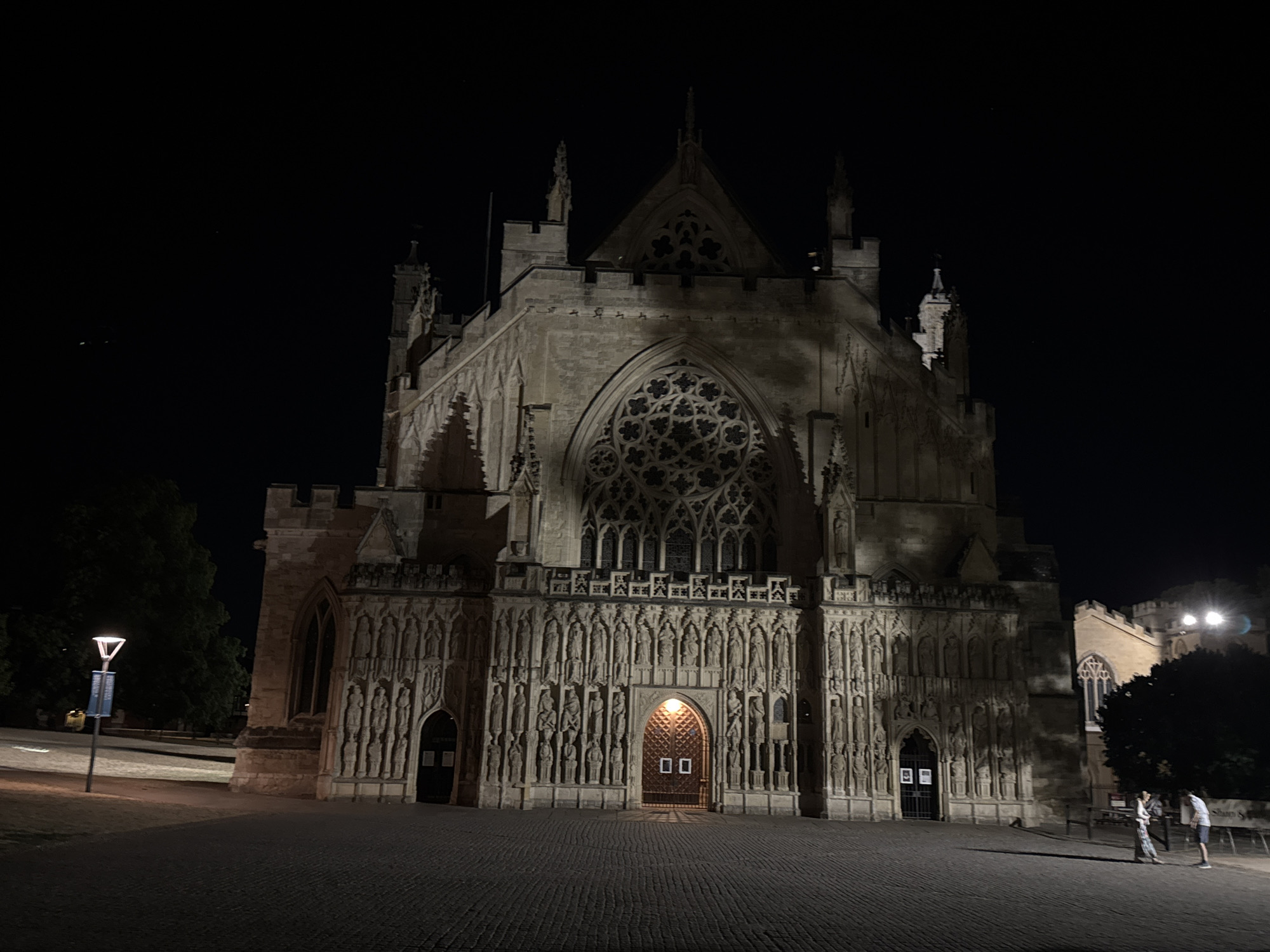 The image shows Exeter Cathedral in Exeter, United Kingdom, at night. The cathedral is a large, imposing Gothic building with intricate stone carvings and a prominent rose window. The facade is illuminated, casting shadows on the paved courtyard in front of it. Two people are standing in the courtyard, facing the cathedral. A street lamp is visible on the left side of the image, and a tree is to the right. The sky is dark, with a few stars visible.