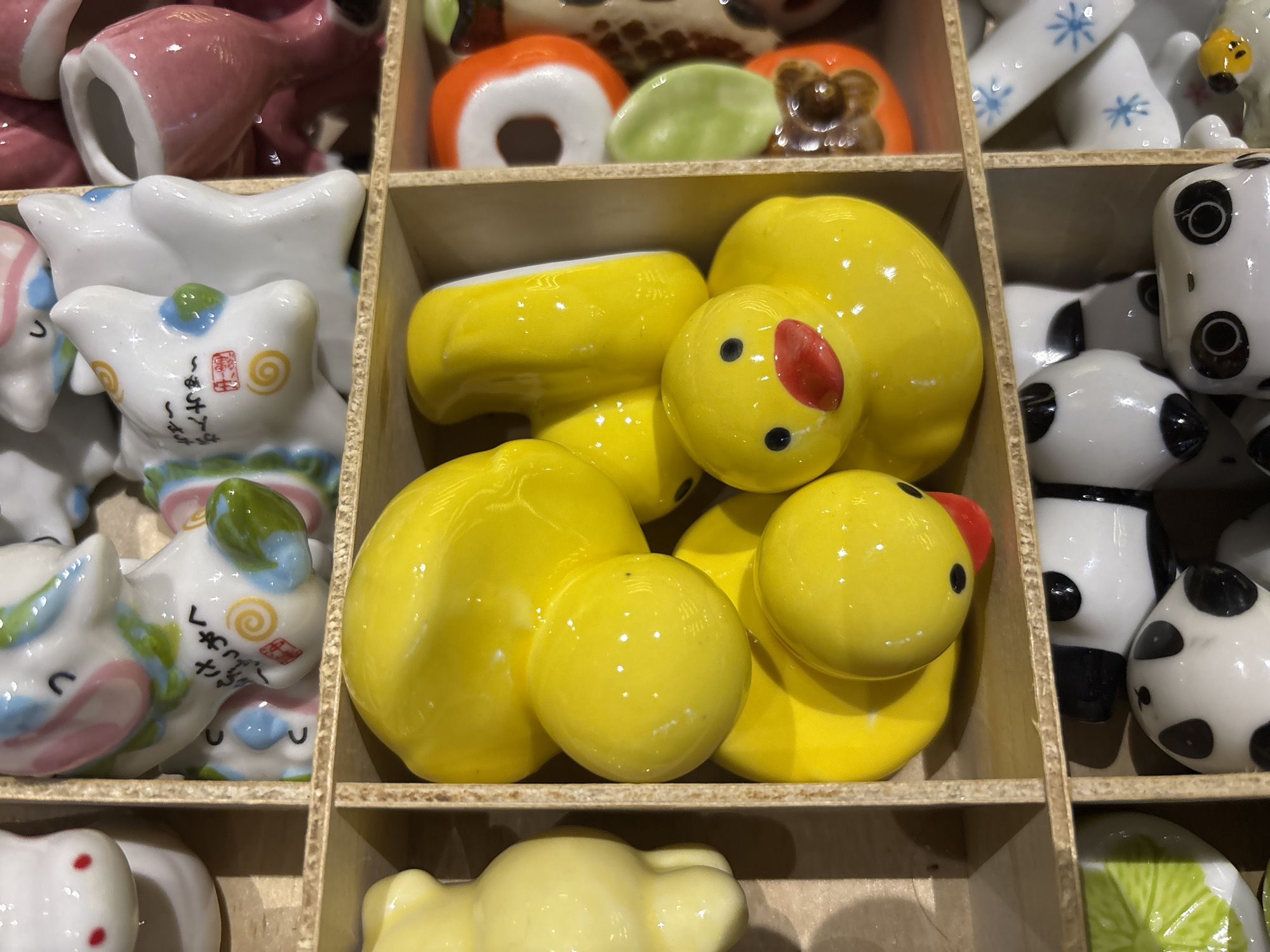 The image shows a grid of small ceramic figurines, possibly souvenirs or decorations, arranged in cubbies. The central focus is a set of yellow duck figurines with red beaks and black eyes, nested within a wooden cubby. Other visible figurines include white and pink unicorn-like creatures with intricate designs, a yellow bear, and various other colorful, abstract shapes. The background suggests a market stall or display area. The image is a close-up shot, with the majority of the frame filled with the ceramic items. There are no people visible in the image, and no specific details indicate the location in Da Lat, Vietnam.