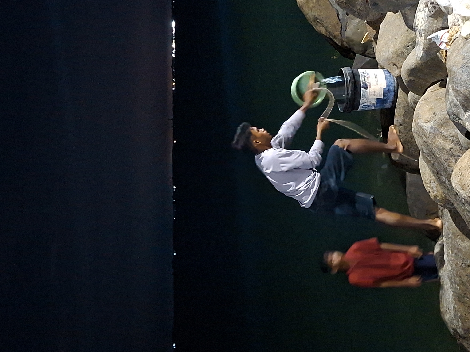 The image depicts a nighttime scene on the rocky shore of the waters in Lhokseumawe, Indonesia. In the foreground, a young man wearing a light-colored long-sleeved shirt and dark shorts is actively pouring water from a green circular object, likely a bowl or lid, into a dark bucket. He is standing on large, rough rocks, with his left foot bare. Next to him, a bucket with a blue and white label is positioned on the rocks. Behind him and to the left, a younger boy in a red t-shirt and dark shorts stands looking forward. The background is dominated by dark water with a faint line of distant lights visible on the horizon, suggesting the presence of a city or settlement across the water. The overall atmosphere is dark and somewhat mysterious, illuminated by the artificial lights of the distant landscape and possibly some ambient light on the subjects.