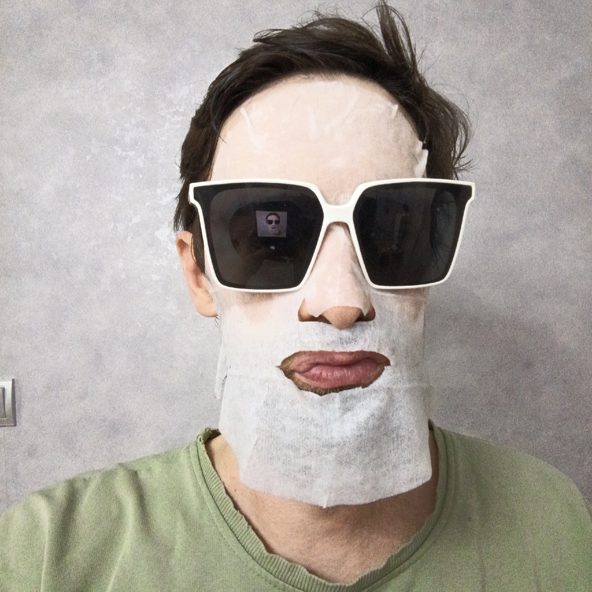 A person wearing a sheet face mask and white-rimmed sunglasses is centered in the frame against a gray wall. The mask has cutouts for the eyes, nose, and mouth. The sunglasses reflect a small image of the person wearing sunglasses. The person is also wearing a light green shirt. A light switch is visible on the left side of the image. There is no verifiable evidence suggesting this image was taken in Марковский, Russia.