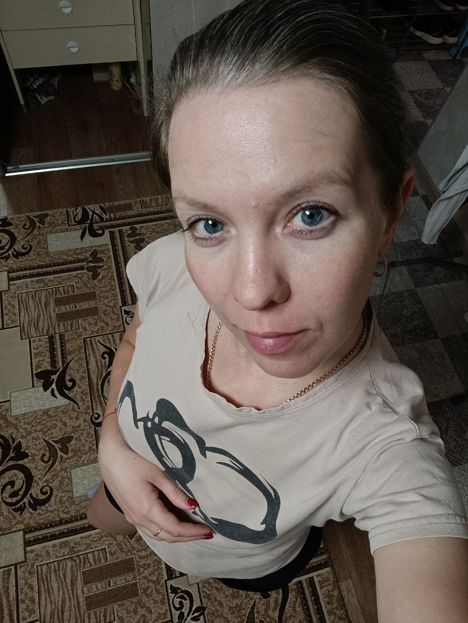 A high-angle selfie depicts a fair-skinned woman with light gray/brown hair pulled back, blue eyes, and light pink lips. She is wearing a beige t-shirt with a black graphic print, a thin gold chain necklace, and small gold hoop earrings. Her left hand is visible, with red nail polish and a gold ring. The background shows a patterned carpet and a cabinet with drawers. The setting may be in Chaikovsky, Russia, as indicated by the prompt, but this is not directly verifiable from the image.