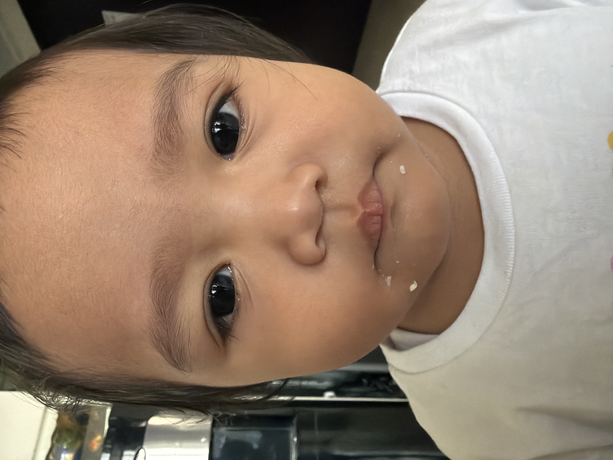 A close-up, head-and-shoulders shot shows a young child, likely a toddler, looking directly at the camera. The child has dark hair, dark eyes, and light brown skin. They are wearing a white t-shirt. There are small crumbs of food on and around the child's mouth. The background is blurred but appears to be indoors, with a dark vertical element on the right and a lighter vertical element with some objects visible on the left. The scene is likely a home environment in Central Jakarta, Indonesia.