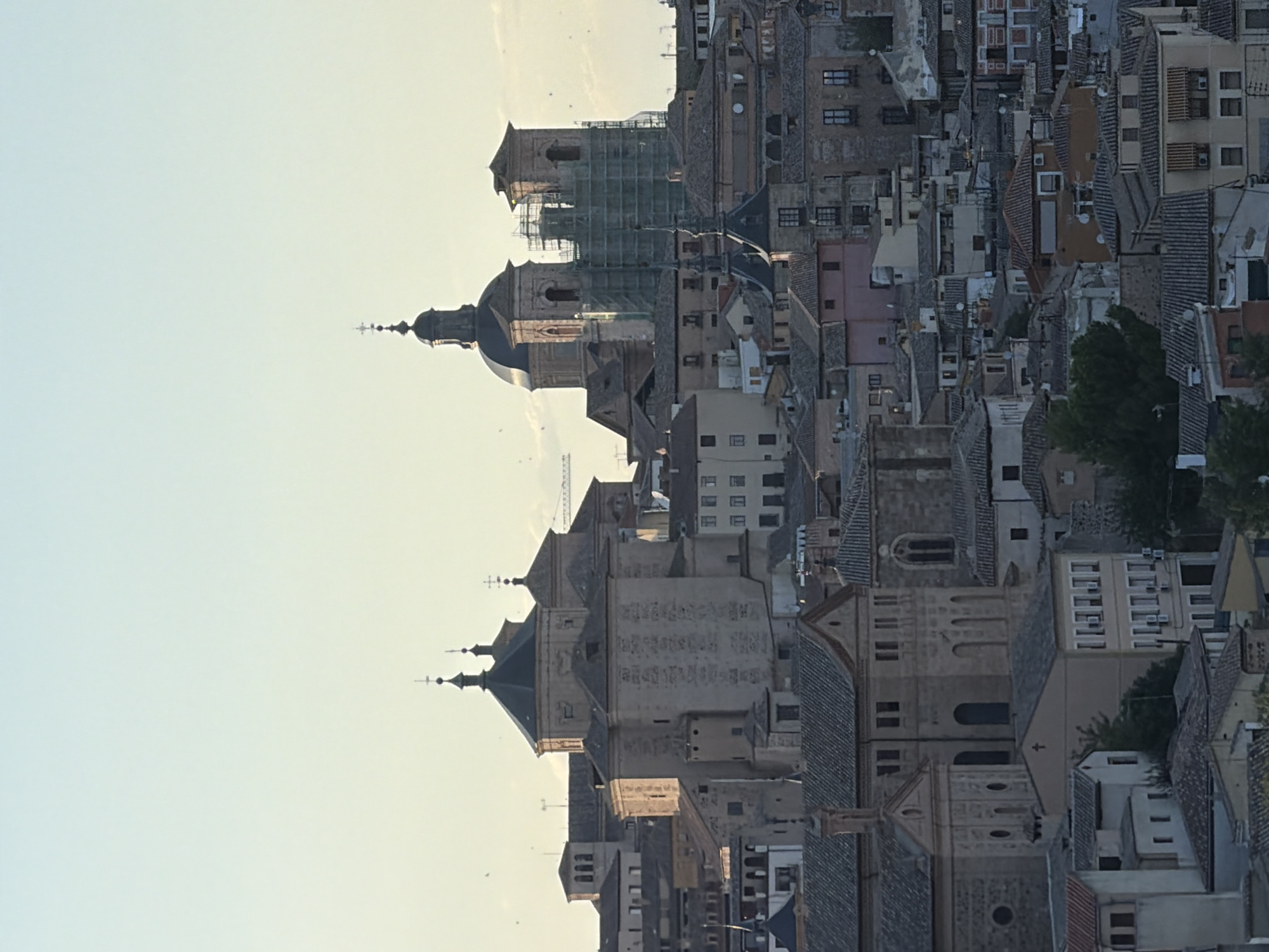 This is a medium-range shot of the city of Toledo, Spain, taken from a high vantage point. The cityscape is dominated by terracotta-tiled roofs and stone buildings, typical of historic European architecture. Two prominent church structures with domes and spires are visible, one of which is partially covered in scaffolding, suggesting ongoing renovation. The sky is a pale, clear blue, indicating daylight, possibly late afternoon or early morning given the soft light. The overall impression is of a densely built, ancient urban landscape bathed in gentle sunlight.