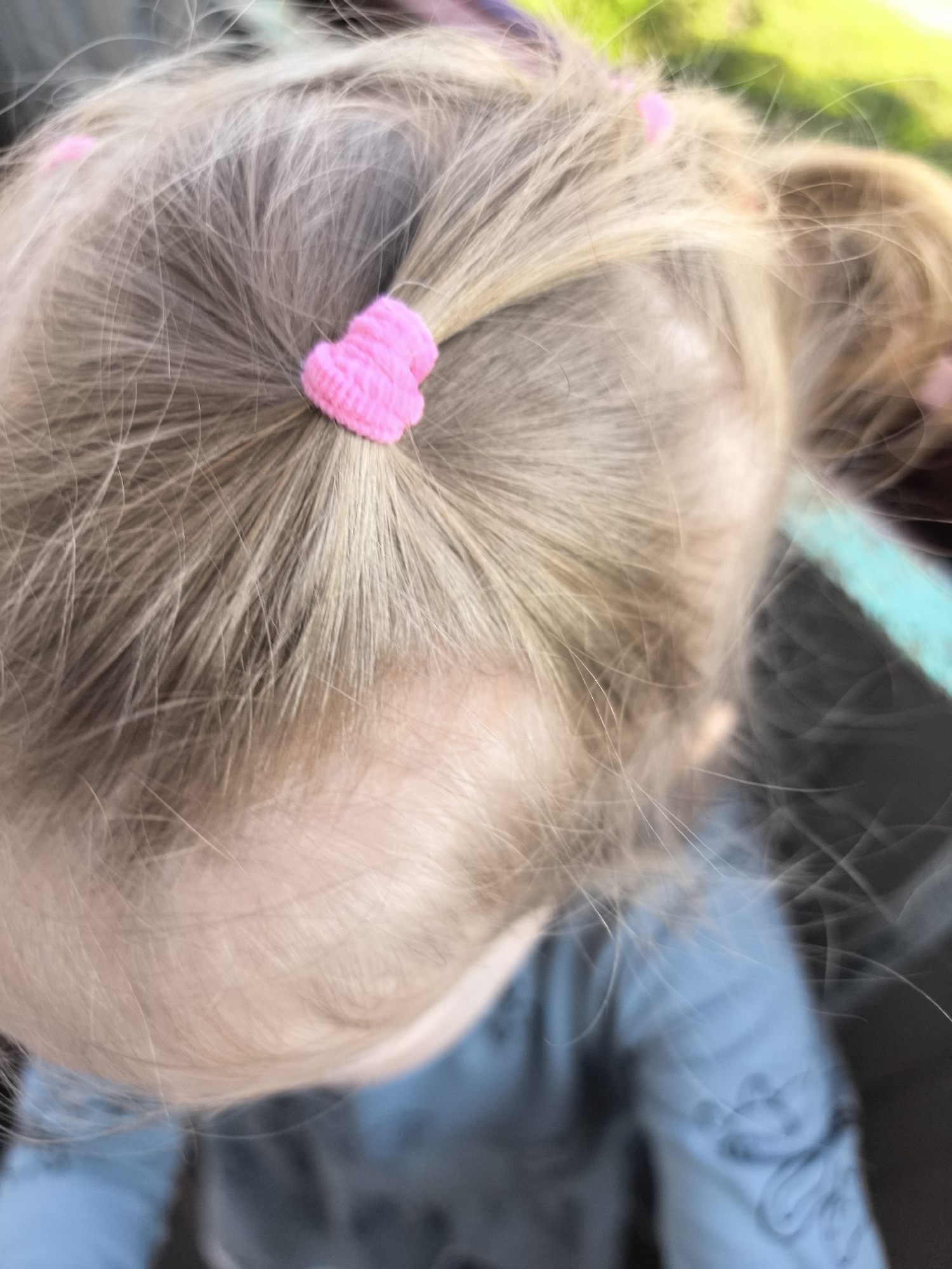 The image is a close-up, overhead shot of the back of a young child's head, focusing on their blonde hair. The hair is styled in pigtails, each secured with a small, bright pink, heart-shaped hair tie. The child is wearing a blue shirt. The background is blurred, showing hints of green foliage and a light blue or teal object. The setting appears to be outdoors, possibly in a park or garden, but no specific city or location details are discernible from the image.