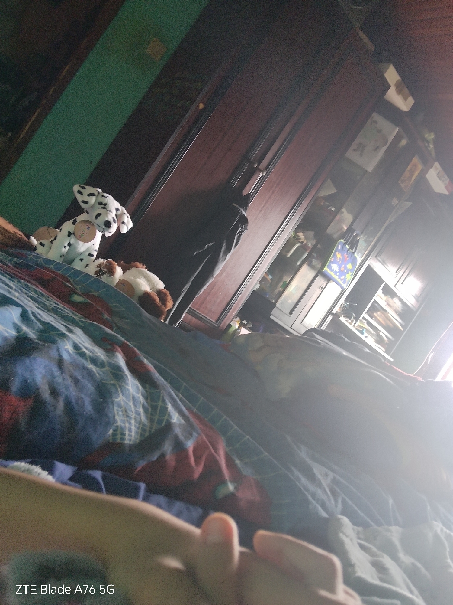 The image depicts an indoor scene, likely a bedroom, with a bed covered in a blue and red patterned comforter. Two plush toys are visible on the bed: a dalmatian toy and a brown and white toy. To the right, a tall, dark wooden wardrobe with glass-fronted doors is partially visible, containing shelves with various items. The overall lighting suggests it might be daytime, with a bright light source creating glare on the right side of the image. A hand is in the foreground, obscuring part of the view. The lower-left corner of the image displays text identifying the device used, "ZTE Blade A76 5G". No specific location in Stara Zagora, Bulgaria is identifiable from the visual information provided.