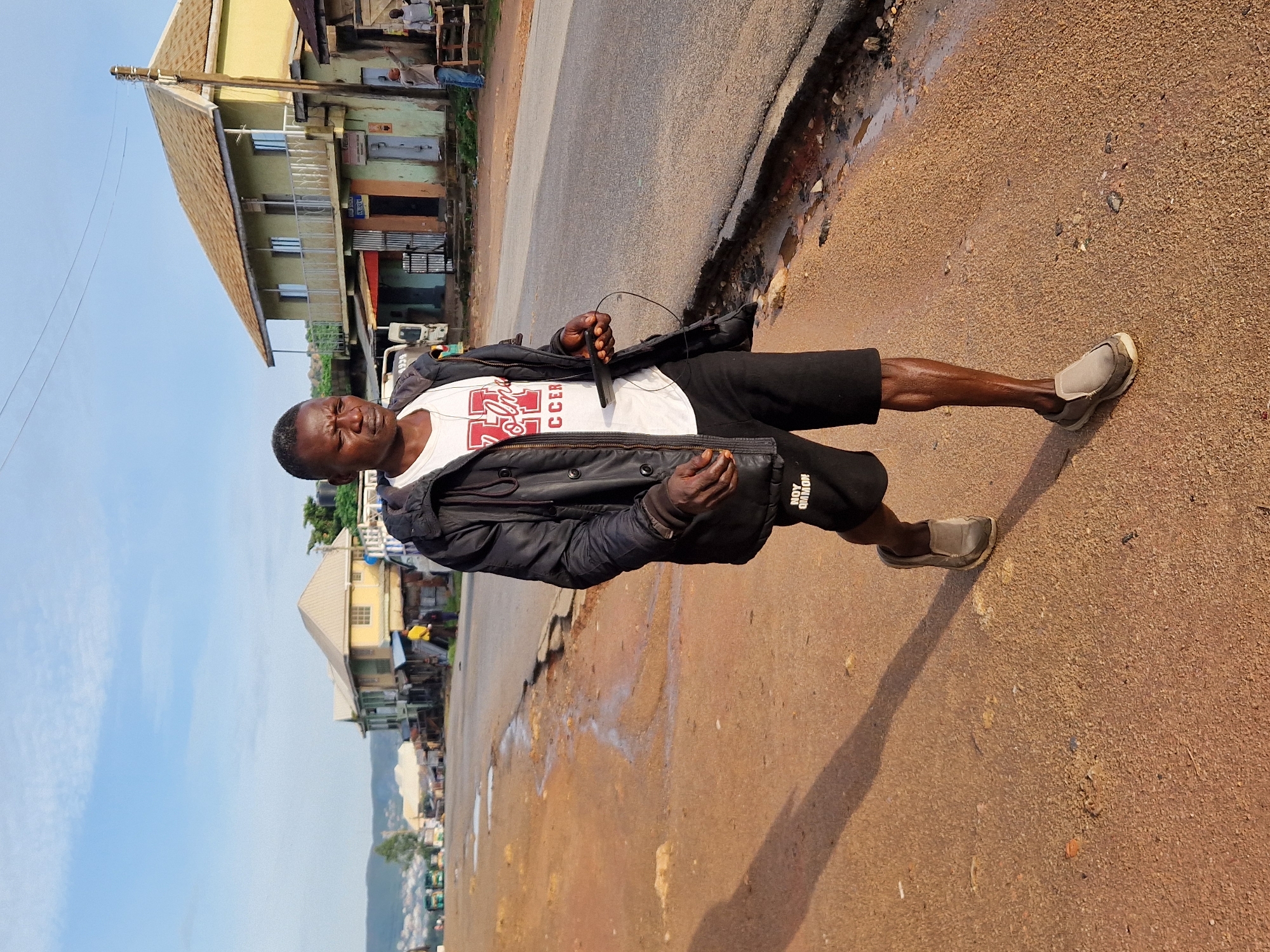 A Black man walks along a dirt road on Rukuba Road, Nigeria. He is wearing a black jacket over a white t-shirt and black shorts. He is holding a black cell phone in his right hand and appears to be listening to music through headphones. The road is unpaved and has puddles of water, indicating recent rain. In the background, there are several buildings, including a two-story building with a green facade and a yellow roof. There are also some vehicles parked along the side of the road. The sky is blue with some scattered clouds. The overall scene suggests a rural or semi-urban environment.