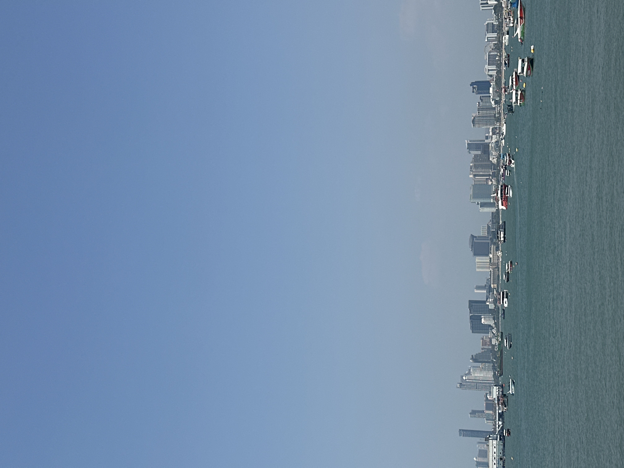 The image shows an aerial view of Pattaya City, Thailand, along the coastline. The right side of the image features the ocean, with numerous boats and vessels visible. The coastline is lined with a dense concentration of buildings, primarily high-rise structures, indicating a developed urban area. The left side of the image is dominated by a clear, blue sky. The overall impression is a panoramic view of a coastal city, likely taken from a high vantage point.
