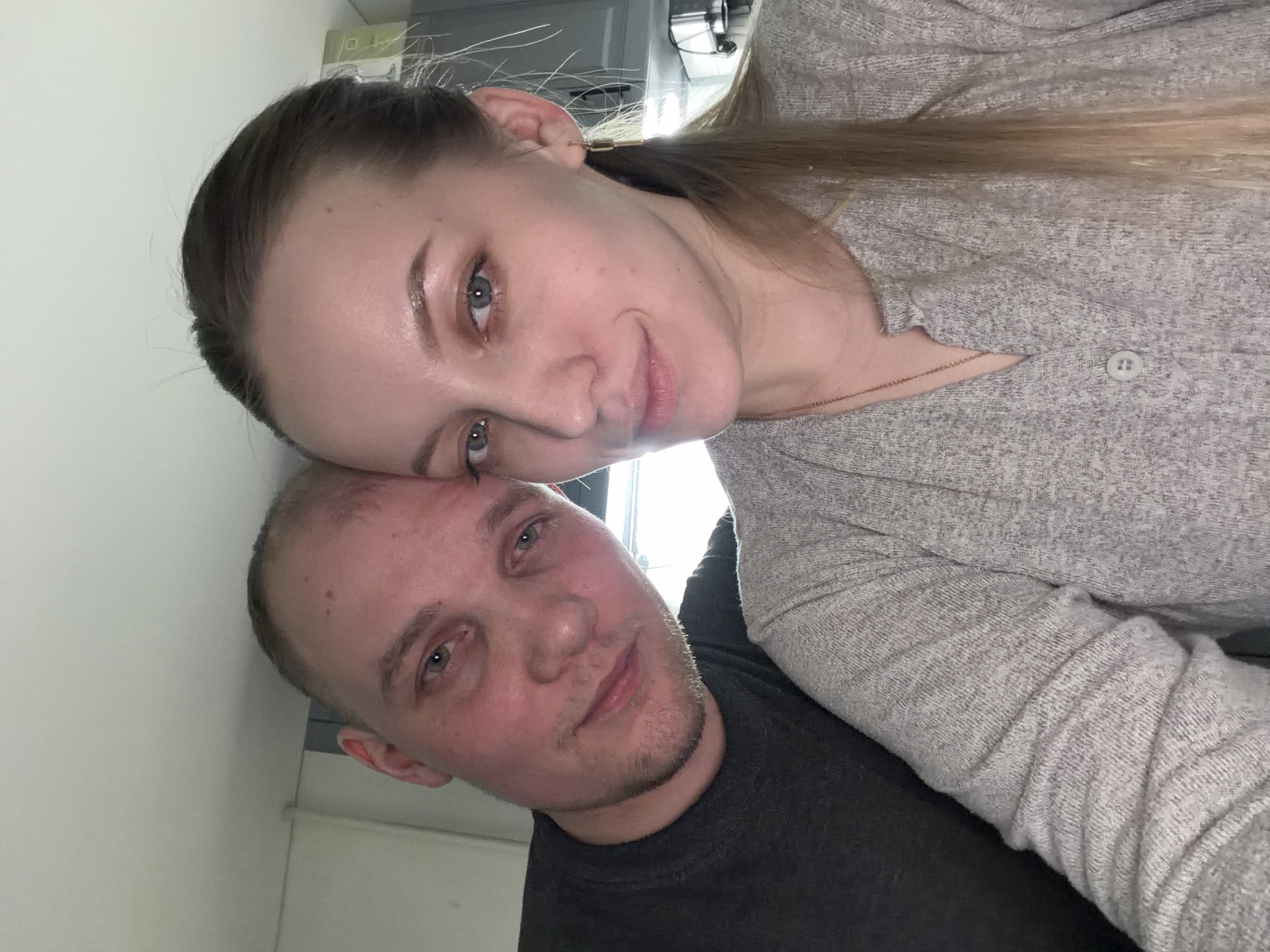 A selfie of a woman and a man. The woman has fair skin, light eyes, and light brown hair pulled back in a ponytail. She is wearing a light grey, button-down shirt. The man has fair skin, light eyes, and short, light brown hair. He is wearing a dark grey t-shirt. The background is a white wall and some kitchen cabinets. The location is not discernible from the image. The image does not contain information regarding a city in Russia.