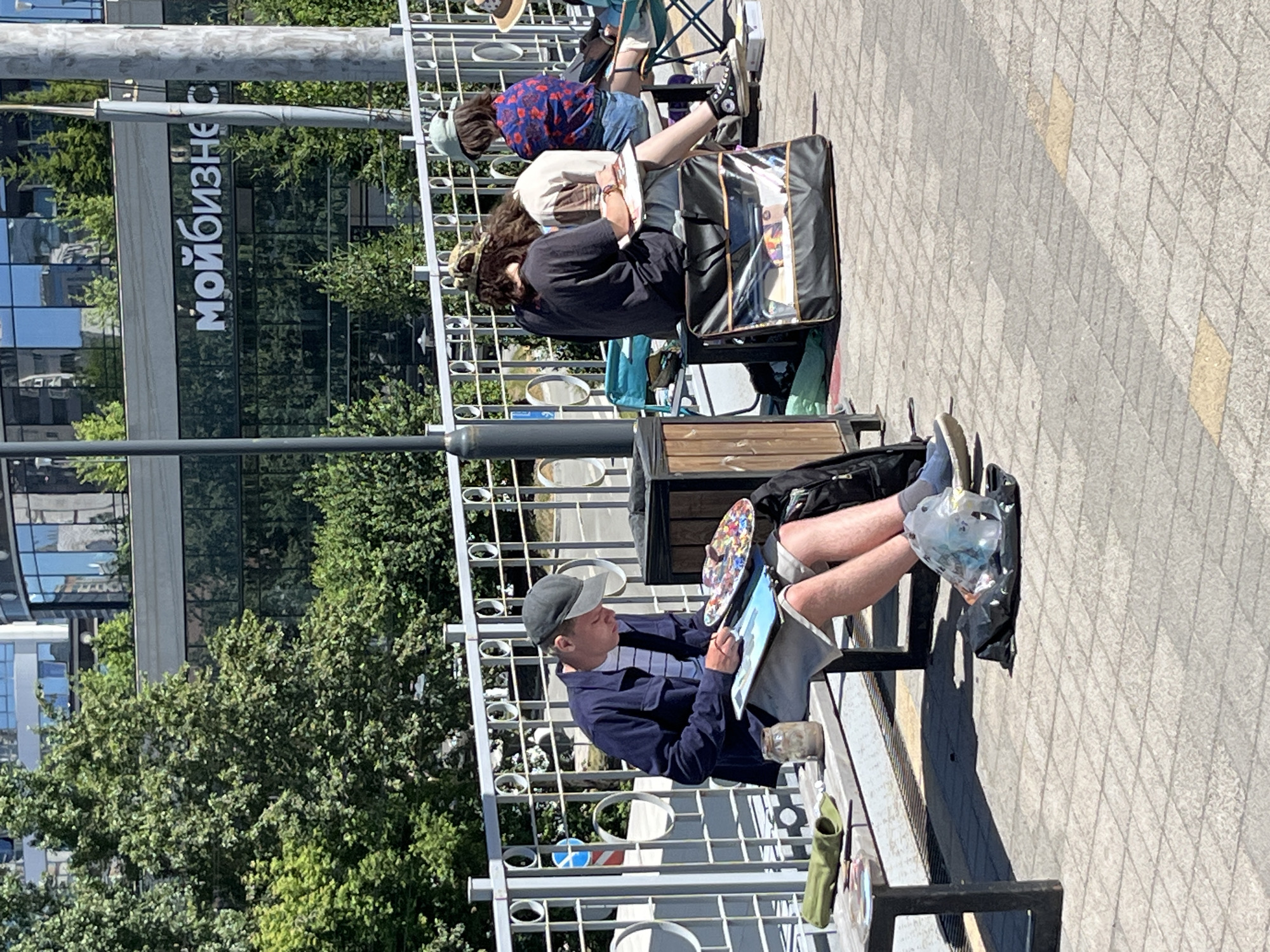 In an urban setting, a group of people are gathered outdoors on a sunny day, engaged in artistic activities. A young man, wearing a dark blue jacket and a grey baseball cap, sits on a metal bench on the left side of the frame. He is actively painting, holding a palette in his right hand and working on a canvas or drawing pad. A jar of water and art supplies are visible on the bench next to him. In the background, behind a white metal railing, is a modern building with a glass facade. Further to the right, two other individuals are also seated, appearing to be painting or drawing. One person, seen from behind, has curly brown hair and wears a black t-shirt, and another individual to their right wears a patterned blue and red shirt. A large black art bag with a transparent compartment is positioned on the ground in front of them. The ground is paved with grey square tiles, and the scene is bordered by green trees and vegetation. The overall impression is one of an outdoor art workshop or sketching session in a public urban space.