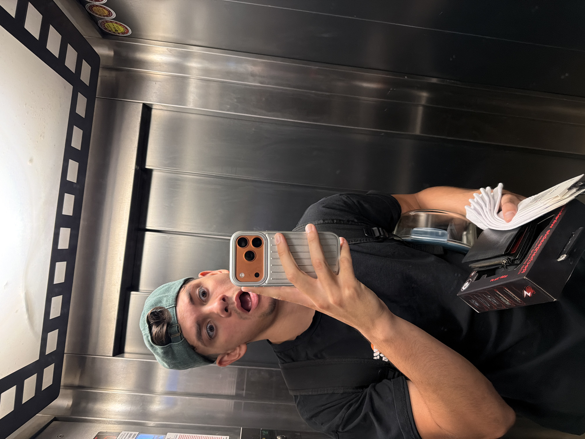 In an elevator with metallic walls, a person wearing a green cap and a black t-shirt takes a selfie. They are holding a smartphone in their right hand, which is angled towards the mirror. The person's mouth is open in an exaggerated expression of surprise or excitement. In their left hand, they are holding a black box with red lettering and a stack of white folded napkins. The elevator ceiling has a decorative pattern resembling a film strip. There are circular stickers on the right wall of the elevator. The overall scene suggests a mundane moment captured in an urban environment, possibly within a building in Blagoevgrad, Bulgaria, given the context of the city.