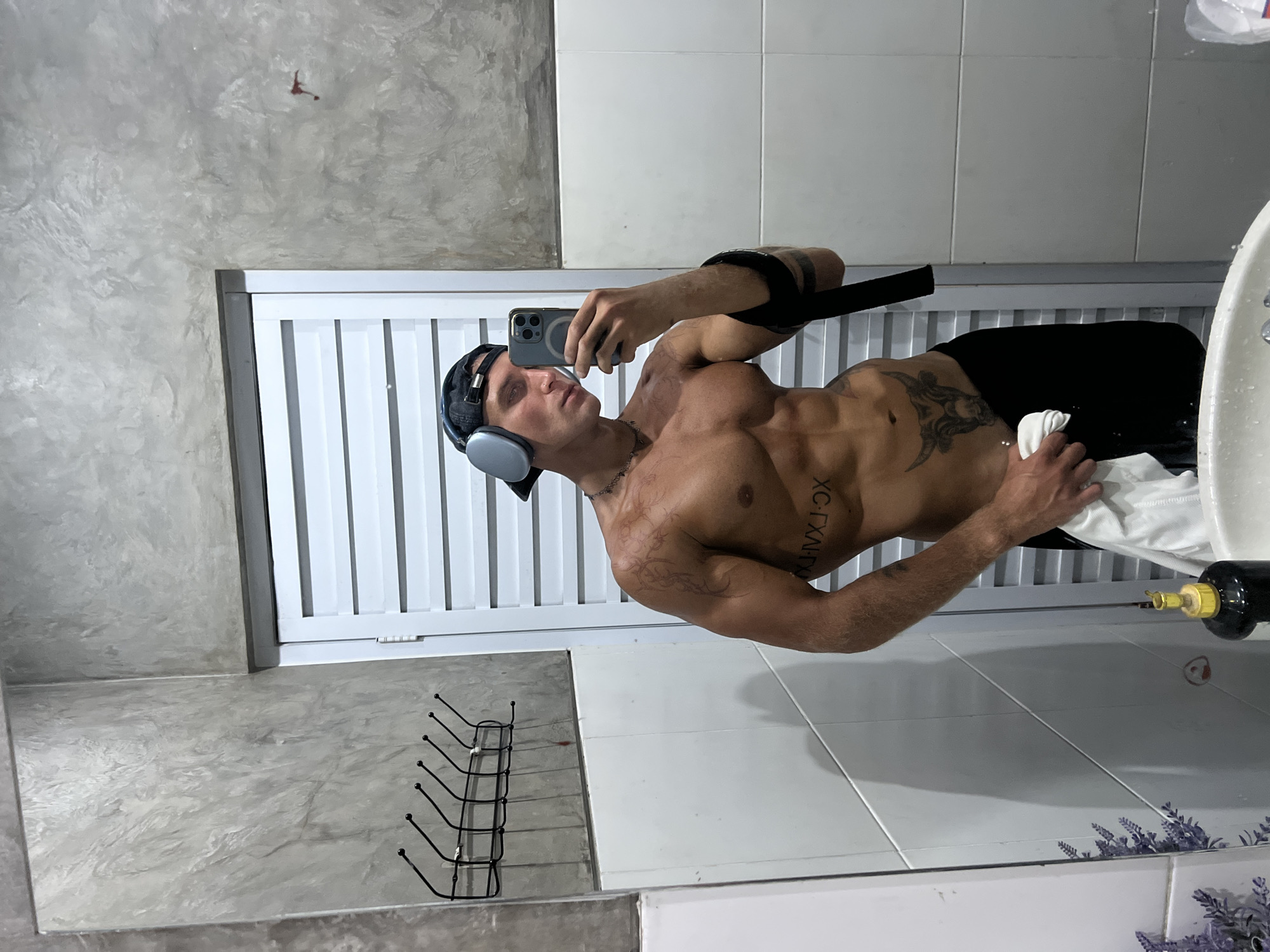 A shirtless, muscular man with tattoos stands in a bathroom, taking a mirror selfie. He is wearing headphones and a baseball cap. His left arm is bent at the elbow, and he is holding a white towel in his right hand. The bathroom has concrete walls and white tiled surfaces, with a row of hooks on the wall. A closed white door with horizontal slats is visible behind him. In the foreground, a sink and a bottle of soap are visible. There is a reflection of the man and the bathroom in the mirror. This location is not identifiable as being in the city of North Kuta, Indonesia.