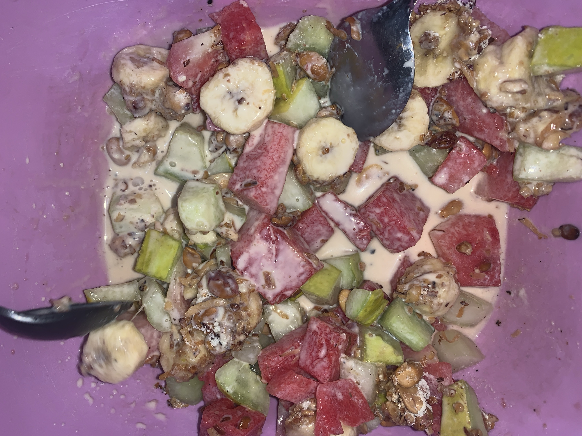 The image provides an overhead close-up view of a fruit salad or dessert served in a purple bowl. The bowl is largely filled with a colorful mixture of diced fruits, mixed with a creamy, pale liquid, which appears to be yogurt, cream, or a milk-based dressing. Granola or nuts are also visible, scattered throughout the mixture.
The primary fruits identifiable are bright red chunks of watermelon and several slices of banana, which are off-white to pale yellow with small dark spots. There are also numerous pieces of a light green fruit, possibly cucumber or green apple, adding another layer of color and texture to the dish. The creamy liquid coats many of the fruit pieces and fills the spaces between them.
A silver spoon is partially visible at different points in the image, submerged into the fruit mixture, suggesting that the dish is either being consumed or has just been prepared. The purple color of the bowl creates a distinct contrast with the varied hues of the fruit.
The lighting is somewhat dim, implying an indoor setting, but there are no specific cues regarding the time of day or weather. There are no people visible in the image, nor is there any discernible text. The image solely focuses on the food item. Based on the provided context, this dish originates from Rafin Par, Nigeria.