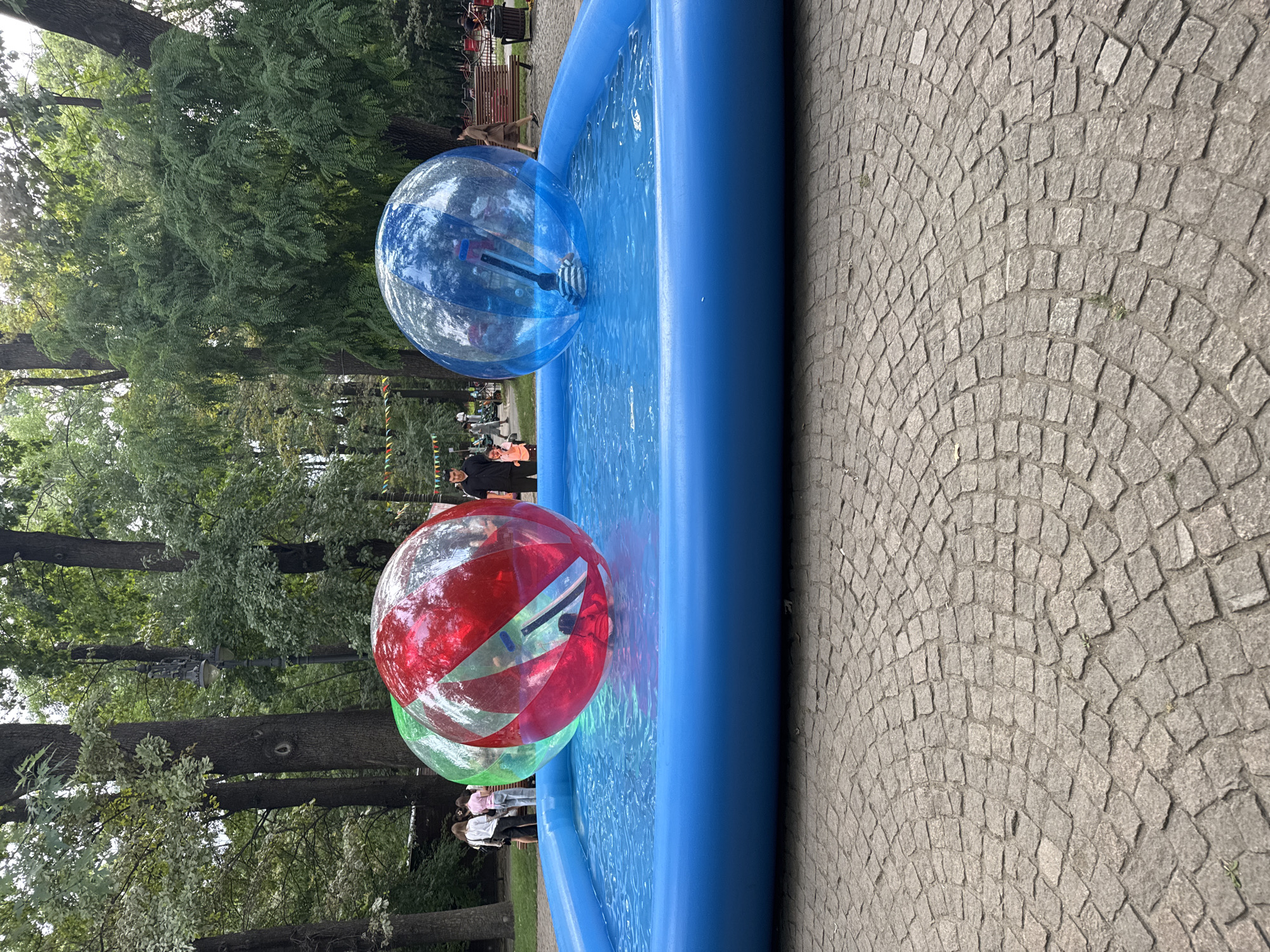 In the city of Almaty, Kazakhstan, at an outdoor event, a blue inflatable pool is filled with water. Two large, transparent inflatable balls, one red and one green, are floating in the pool, each with a person inside. A third, blue inflatable ball is also in the pool with a person inside. People are visible in the background, some standing and watching. The environment suggests a park or public recreational area, with trees and cobblestone pavement in the foreground. The event appears to be a recreational activity for visitors.