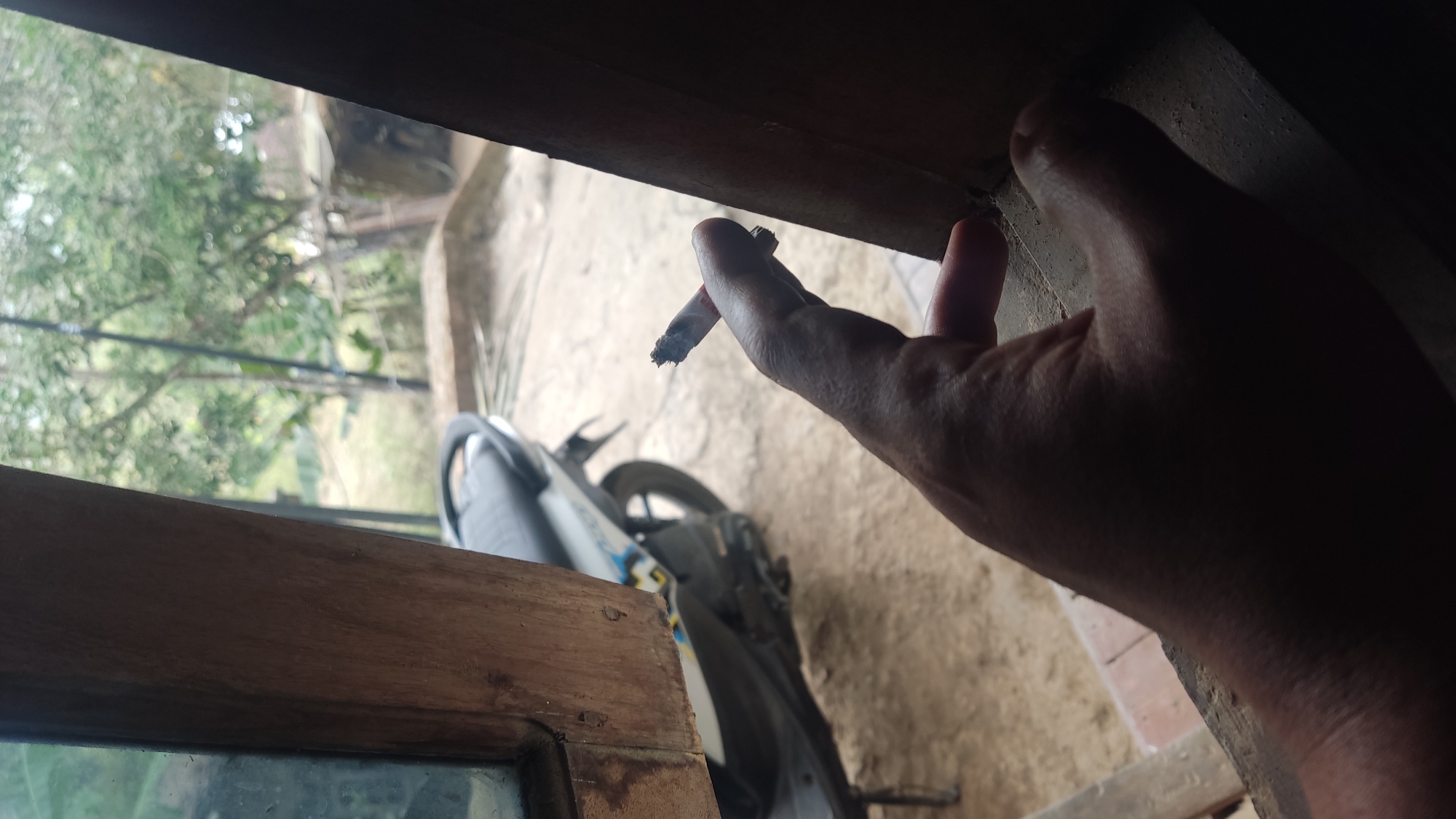 This media file captures a close-up of a person's hand holding a lit cigarette, with the foreground dominated by a wooden frame, possibly a window or doorway. The hand is reaching towards a dark, wooden surface, while the cigarette dangles, emitting a thin trail of smoke. In the blurred background, a motorcycle is visible, suggesting an outdoor or semi-outdoor setting. Green foliage and a light-colored ground, possibly dirt or concrete, are also discernible. The overall lighting suggests it might be daytime, though the specific time is not clearly indicated. The image appears to be taken from a low angle, looking upwards towards the hand and cigarette. There is no visible text in the media file. The scene evokes a casual, perhaps contemplative moment, given the act of smoking. The location context provided, Ngadiharjo, Indonesia, adds to the understanding of the cultural and environmental setting.