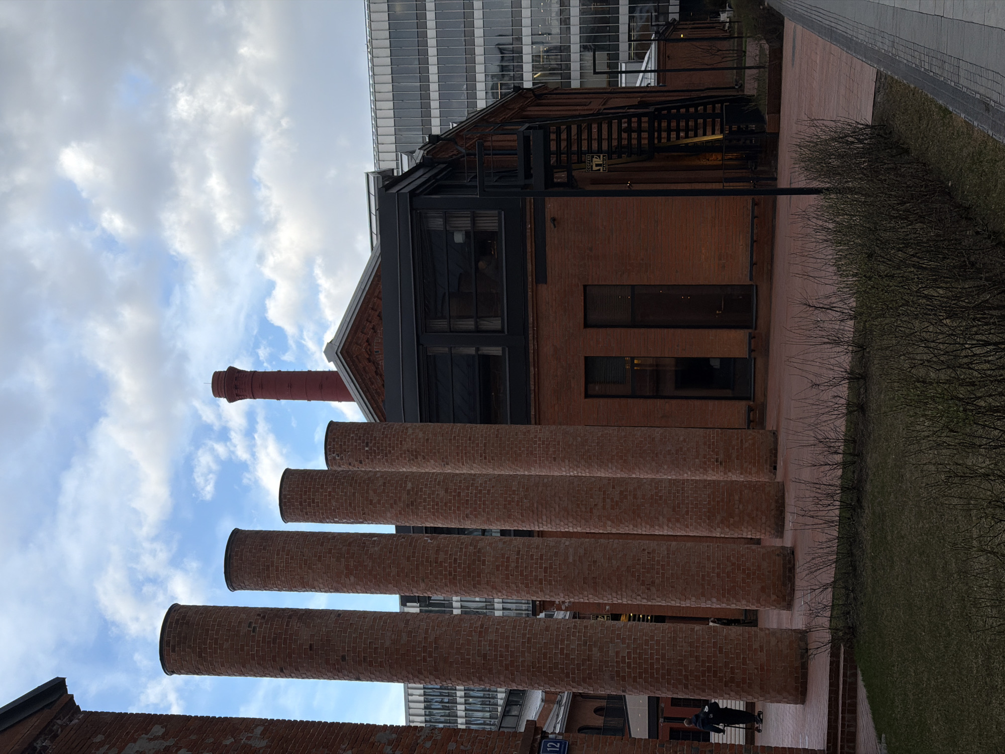 The image captures a scene in Moscow, Russia, specifically the Danilovsky Market area. The prominent features are four cylindrical brick columns extending upward, seemingly remnants of an industrial structure. Behind these, a brick building with a gabled roof and modern glass additions is visible, with a taller glass-clad building in the background. To the right of the brick building, there is a patch of grass and a paved walkway. A person is visible in the lower right corner of the image walking, and what appears to be a small street sign is visible on a brick wall. The sky is partly cloudy.