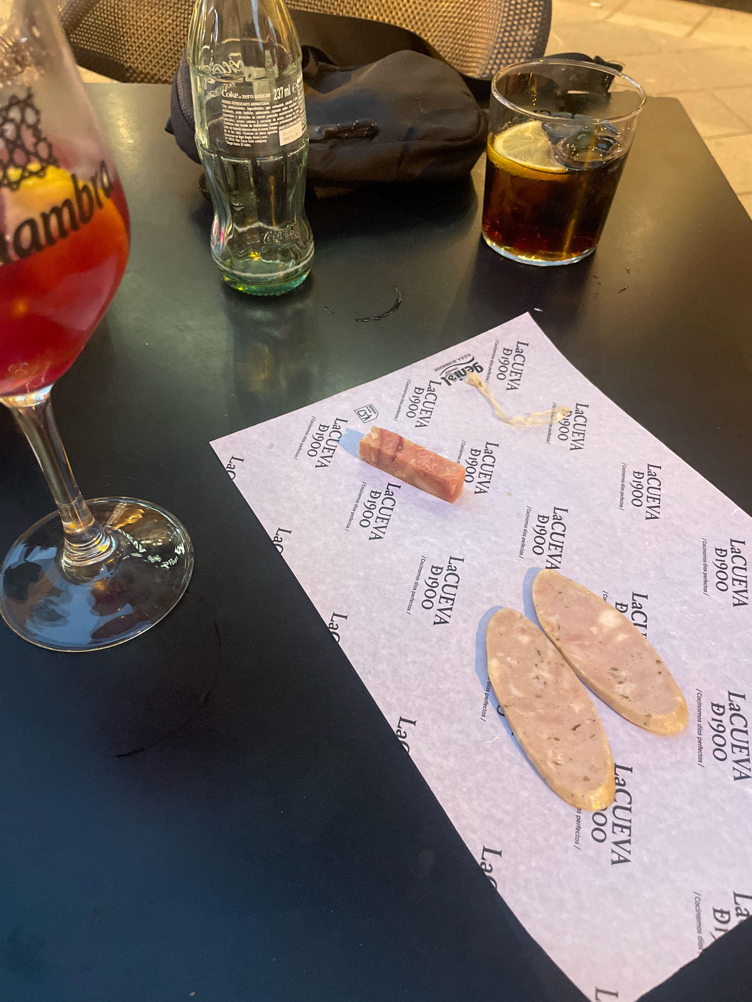 The image displays a close-up view of a dark, possibly black, table at an outdoor cafe. In the foreground, a stemmed glass contains a red-colored beverage with ice and a slice of lemon. To the right of the glass, a transparent glass holds a dark, carbonated beverage with a slice of lemon and ice. Beside these drinks, a white paper liner, printed with the text "LA CUEVA D1900" and "/Cocinamos días perfectos/", is partially visible. On this liner, two oval-shaped slices of processed meat, likely sausage or pâté, and a small rectangular piece of cured meat are arranged. In the background, a glass bottle of Coca-Cola Zero, with its label mostly obscured, is seen next to a black bag. The overall setting suggests a casual dining or drinking experience in a cafe. The presence of "LA CUEVA D1900" on the paper liner strongly suggests the location is at a establishment named "La Cueva D1900". A search reveals that "La Cueva 1900" is a popular tapas bar located in Granada, Spain, known for its traditional Spanish cuisine and atmosphere.