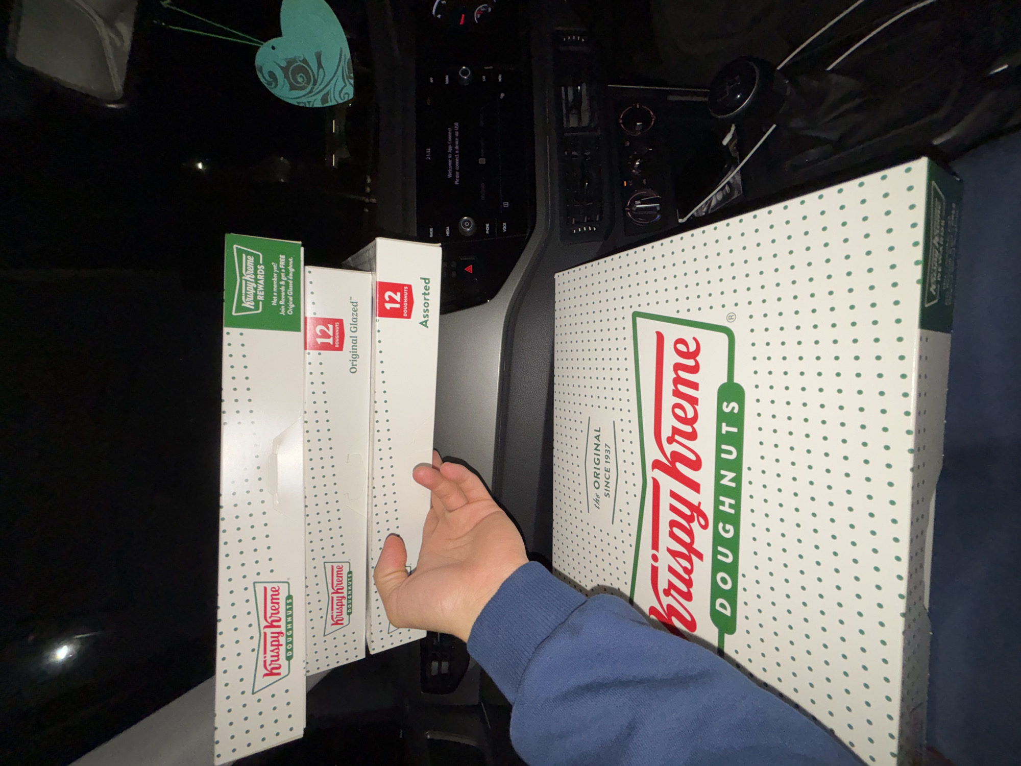 The image shows the interior of a vehicle, likely a car, at night. On the center console, four boxes of Krispy Kreme donuts are stacked. From left to right, the boxes are labeled "Krispy Kreme Rewards," "Original Glazed 12," and "Assorted." A hand is partially visible, touching the assorted box. The fourth box to the right is labelled "Krispy Kreme Doughnuts."
The car's dashboard and center console are visible, showing a display screen and various controls. A heart-shaped object hangs from the rearview mirror. The gear stick is also visible.