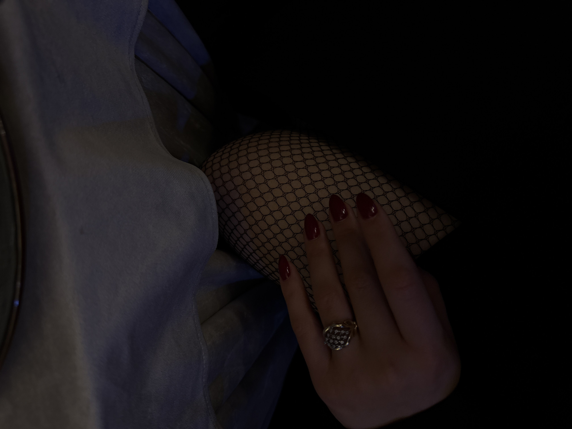 A dark-lit image presents a right hand adorned with red nail polish and a floral-patterned ring resting on a leg covered in black fishnet stockings. To the left is a light blue denim fabric. The background is predominantly dark. There is no indication of Yerevan, Armenia.