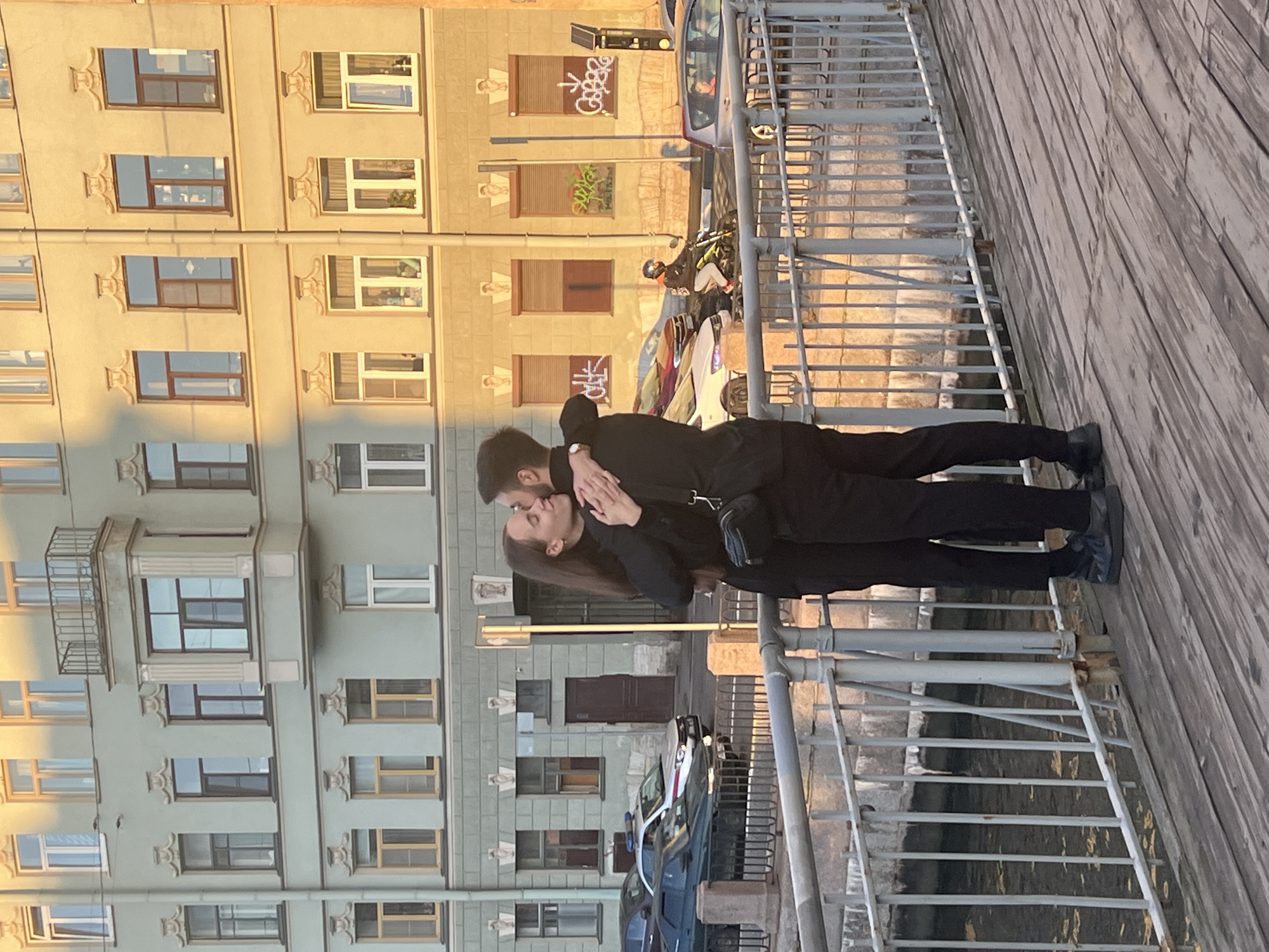 This media file captures a tender moment between a couple embracing on a wooden walkway overlooking a canal in Saint Petersburg, Russia. The couple, positioned in the foreground, are passionately kissing, their bodies closely intertwined. The woman has dark hair that flows down her back, and she wears a black top. The man, also in black, has his arms around her.
Behind them, a metal railing and staircase lead down to the water. The setting is urban, with buildings in the background displaying a mix of architectural styles. One building is light-colored with numerous windows and decorative architectural elements, suggesting an older, historic facade. Another building, to the left, is painted a pale blue-green and also features multiple windows, some with balconies. The sun casts a warm, golden light, indicating it is likely late afternoon or early evening. The shadows are long, further supporting this inference.
Cars and a motorcycle are visible on a street further back, suggesting an active urban environment. Graffiti adorns some of the shutters on the buildings, adding a touch of contemporary street art to the historic backdrop. The overall mood is romantic and intimate, set against the backdrop of a picturesque Saint Petersburg cityscape.