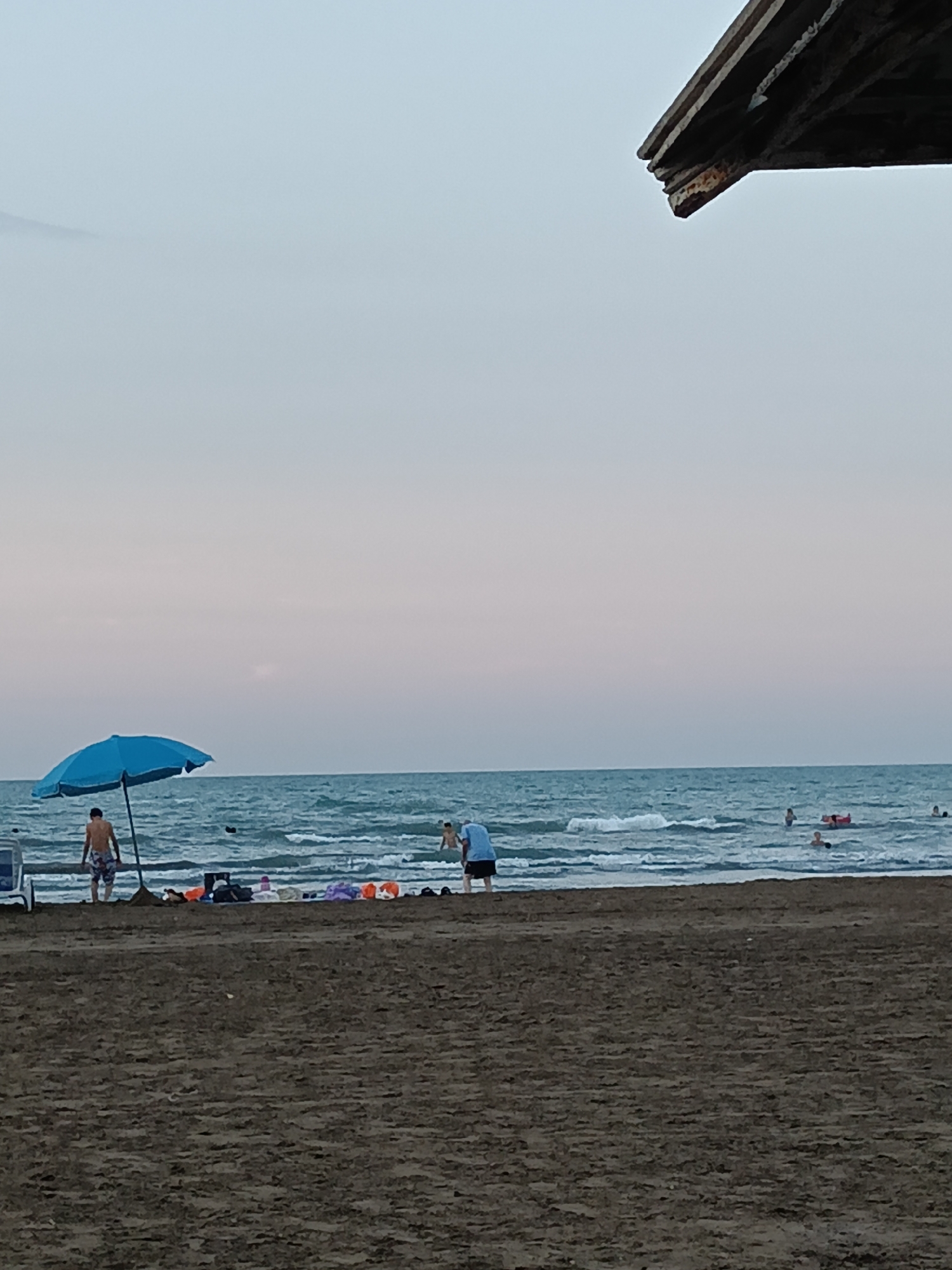 The image captures a sandy beach scene at what appears to be dusk. The sky is a gradient of soft blues and pinks. In the foreground, the dark, wet sand shows texture from ripples. The sea occupies the middle ground, with moderate waves lapping at the shore. A bright blue umbrella stands on the beach near the water's edge, with a person standing nearby wearing patterned shorts. Further into the sea, several people are visible, some swimming or wading, and one person is on an inflatable ring. To the right of the umbrella, a collection of beach items, including bags and towels in various colors, are laid out on the sand. A portion of a wooden structure, likely part of a beach shelter or cafe, is visible in the upper right corner of the frame. The overall atmosphere is relaxed and indicative of a typical beach day.
