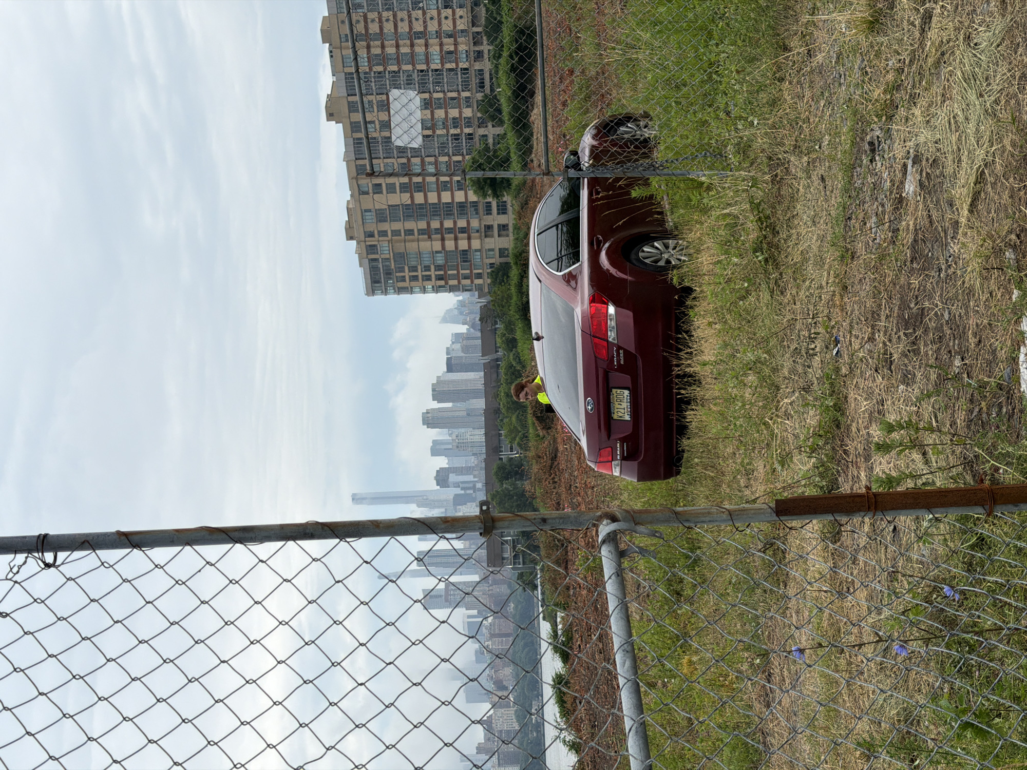 In North Bergen, New Jersey, USA, a red Subaru Legacy sedan is parked in an overgrown lot. The car is facing right, with its rear end visible. A person wearing a yellow safety vest stands near the car. In the background, a cityscape with numerous tall buildings and a river is visible under a cloudy sky. A chain-link fence with rusted poles runs through the foreground of the image, partially obscuring the scene. The ground is covered with dry grass and some small blue wildflowers.